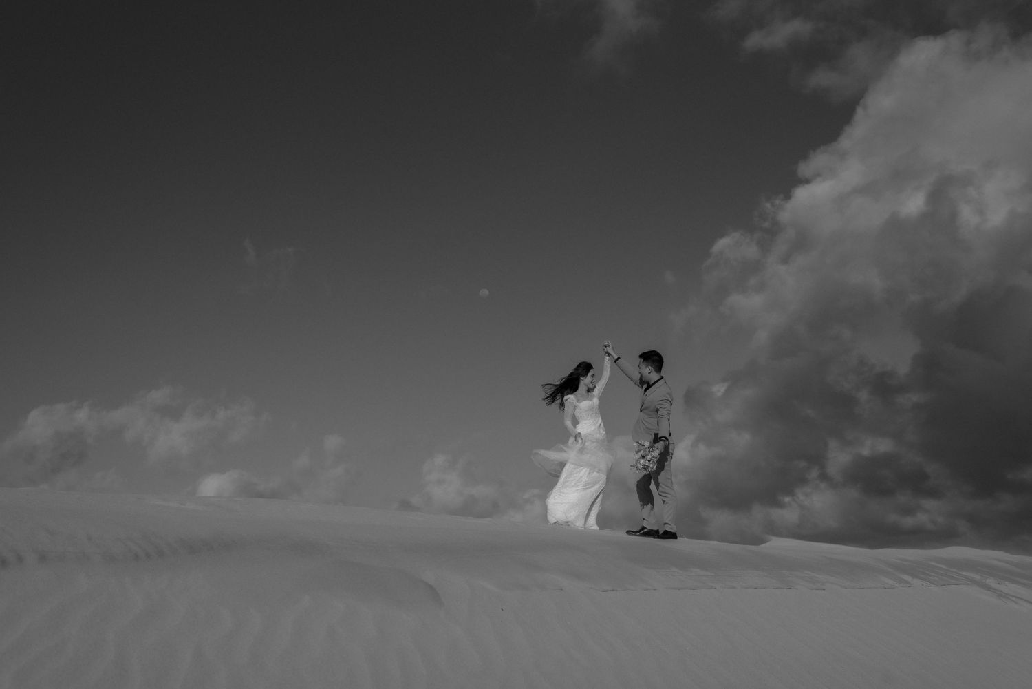 A couple in white wedding attire walks together on a sandy desert dune under dramatic clouds in black and white.