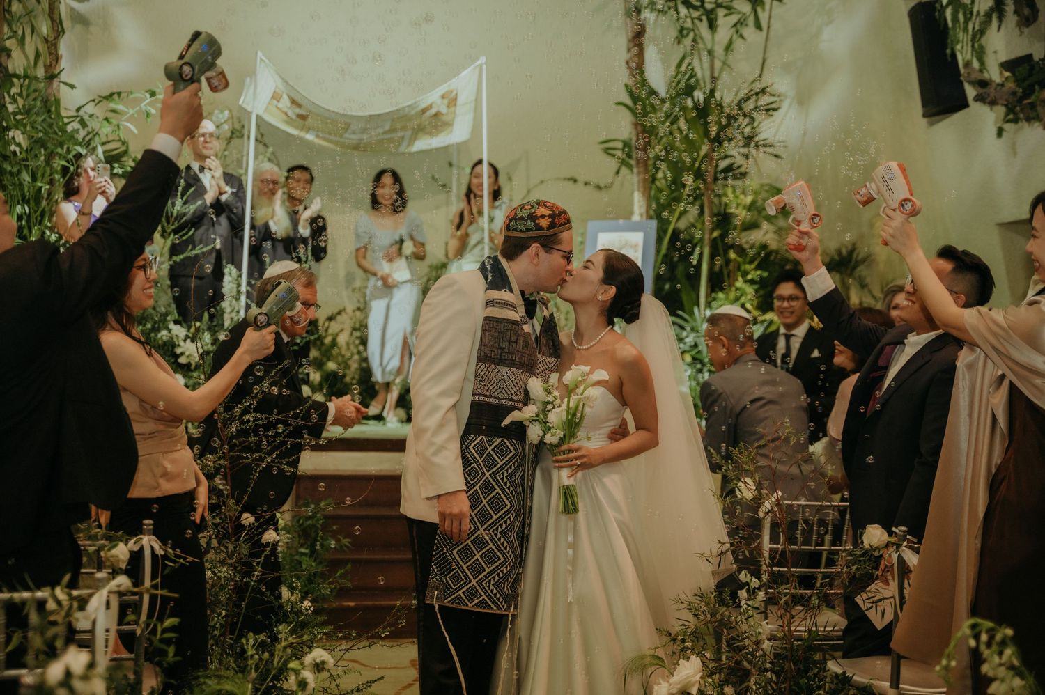 Guests celebrate joyfully during a wedding ceremony in a greenery-filled indoor venue.