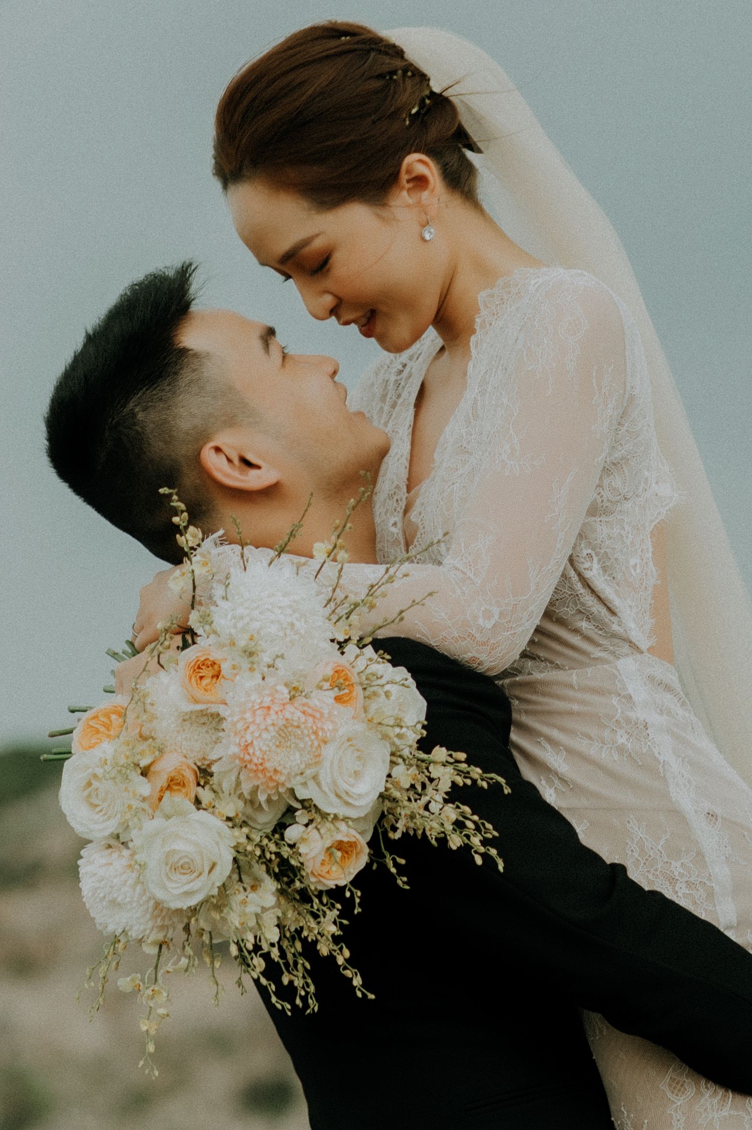 A romantic wedding photo shows a couple embracing while holding a beautiful bouquet of white and peach roses and delicate florals.