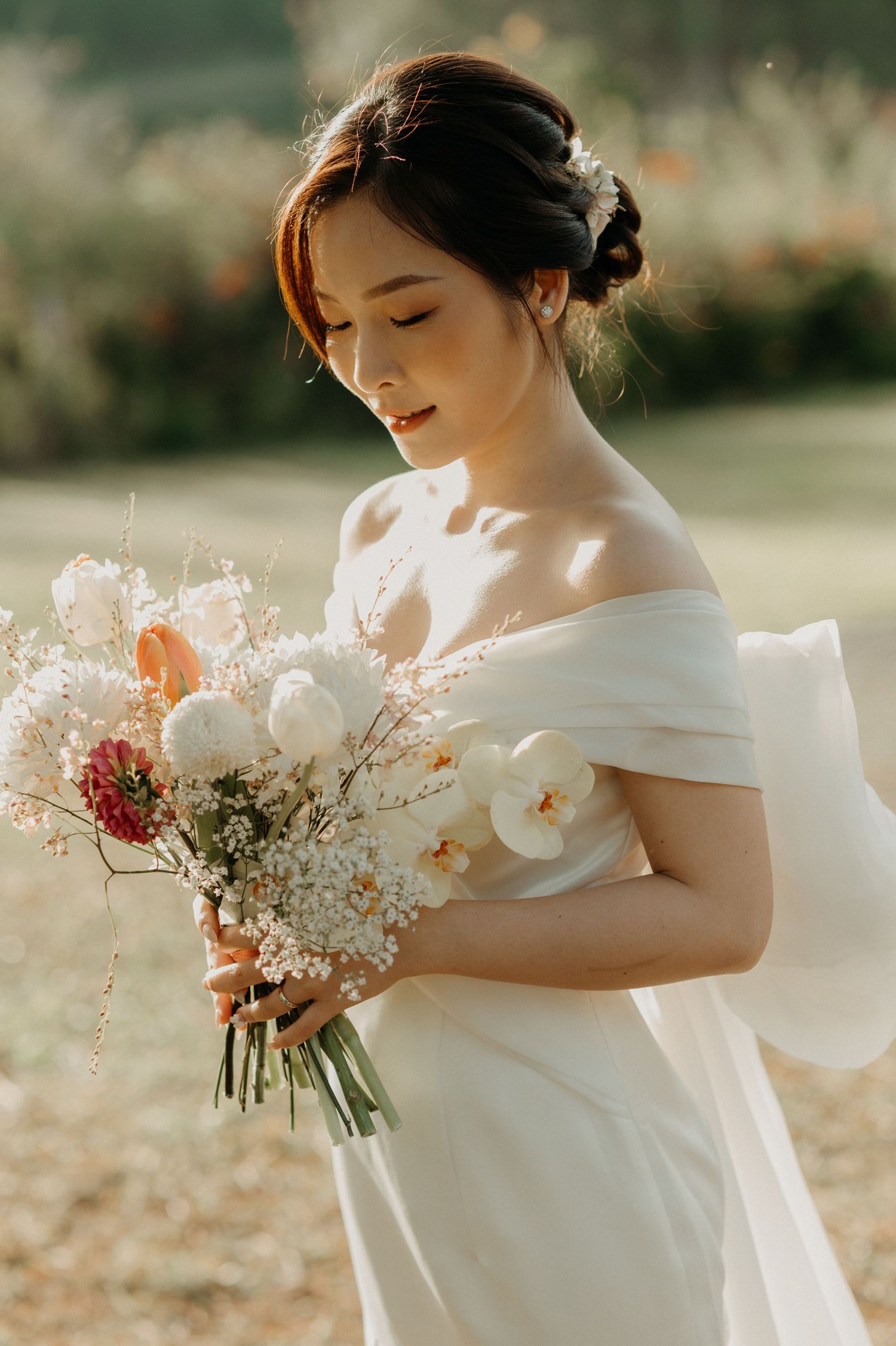 A bride in an elegant off-shoulder white dress holds a delicate bouquet of white and pink flowers in warm sunlit setting.