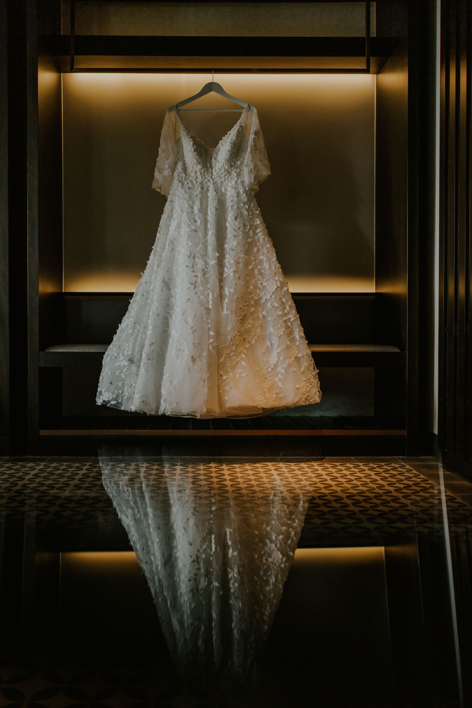 Elegant white lace wedding dress hanging in illuminated display case with reflection.