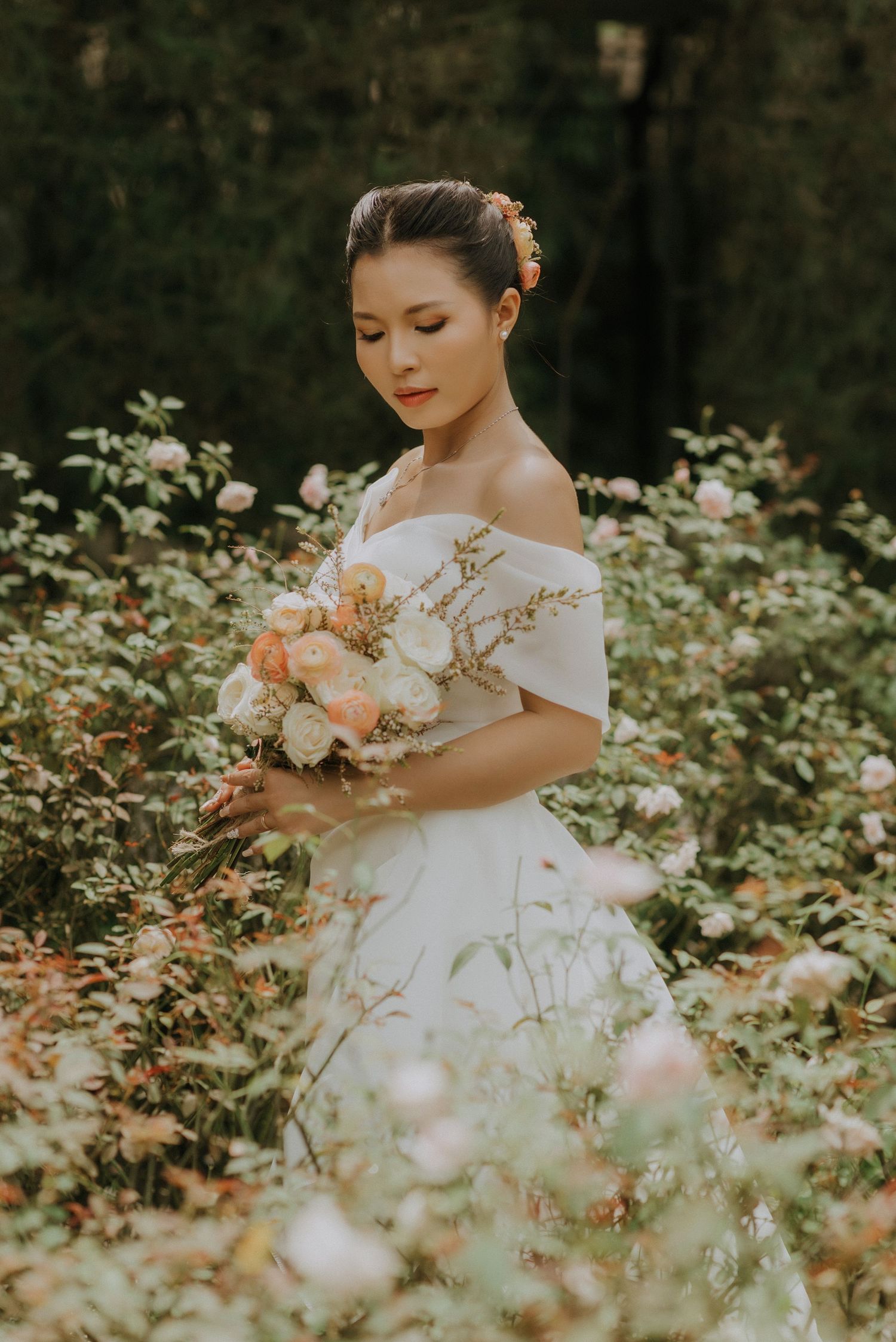 An off-shoulder white wedding dress stands out against lush garden greenery while holding a peach and white floral bouquet.