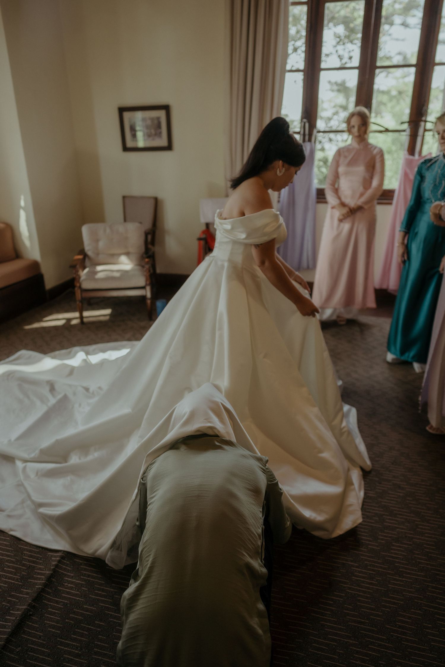 An elegant white wedding dress being fitted in a room with soft natural lighting and pink accents.