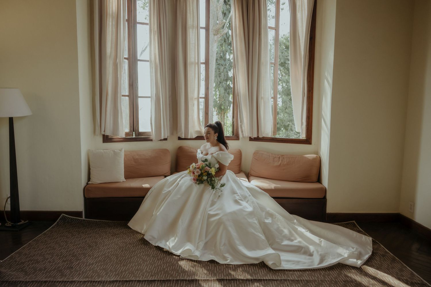 A bride in a flowing white wedding gown sits elegantly on a pink sofa by large windows in a corner room.