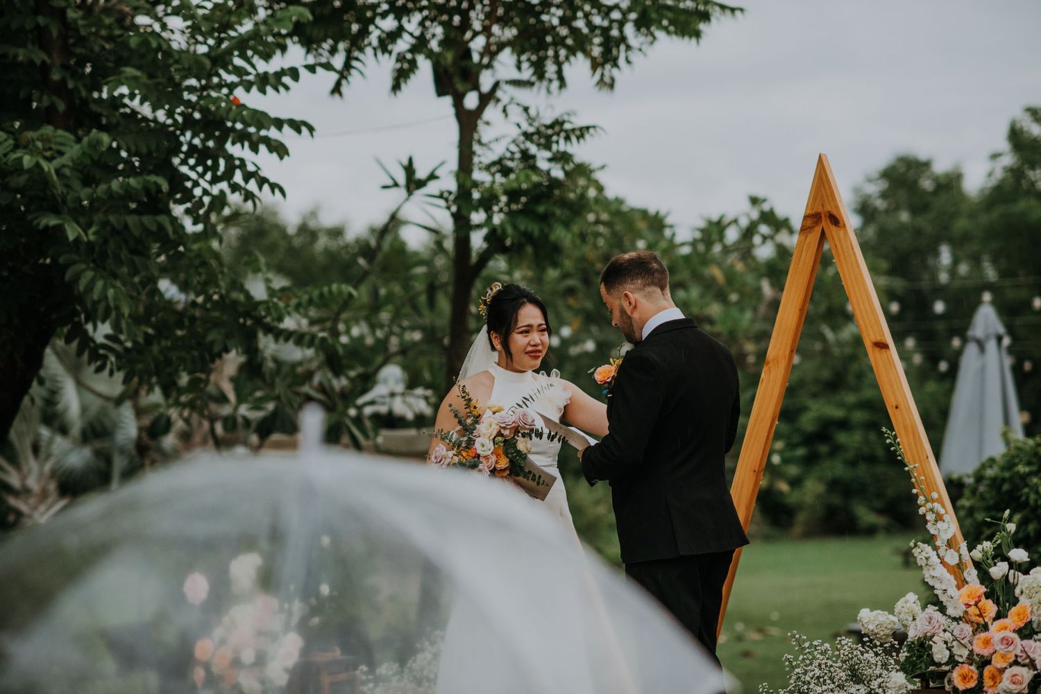 A wedding ceremony takes place under a triangular arch in a misty garden setting.
