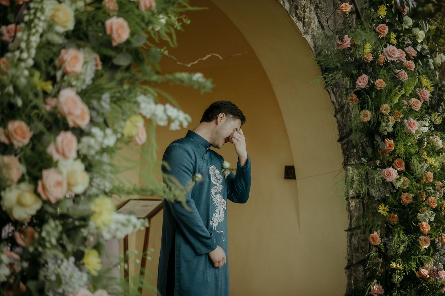 A person in a blue suit stands beneath an elegant archway adorned with blooming pink roses and flowers.