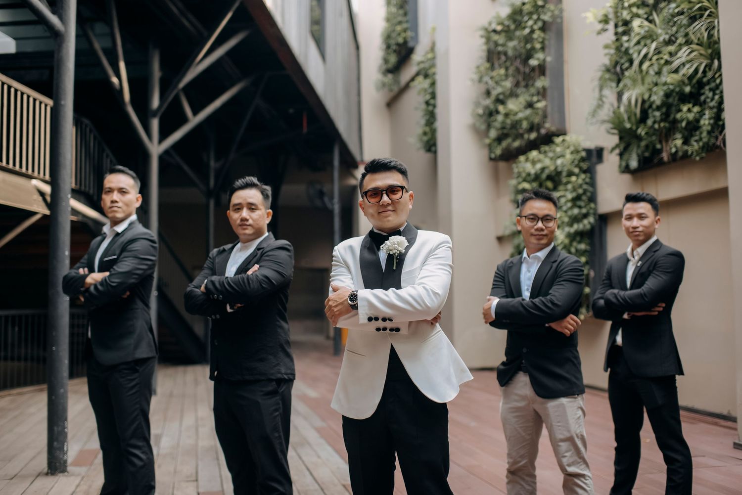 Groomsmen pose confidently in suits with center figure wearing white jacket in courtyard setting.