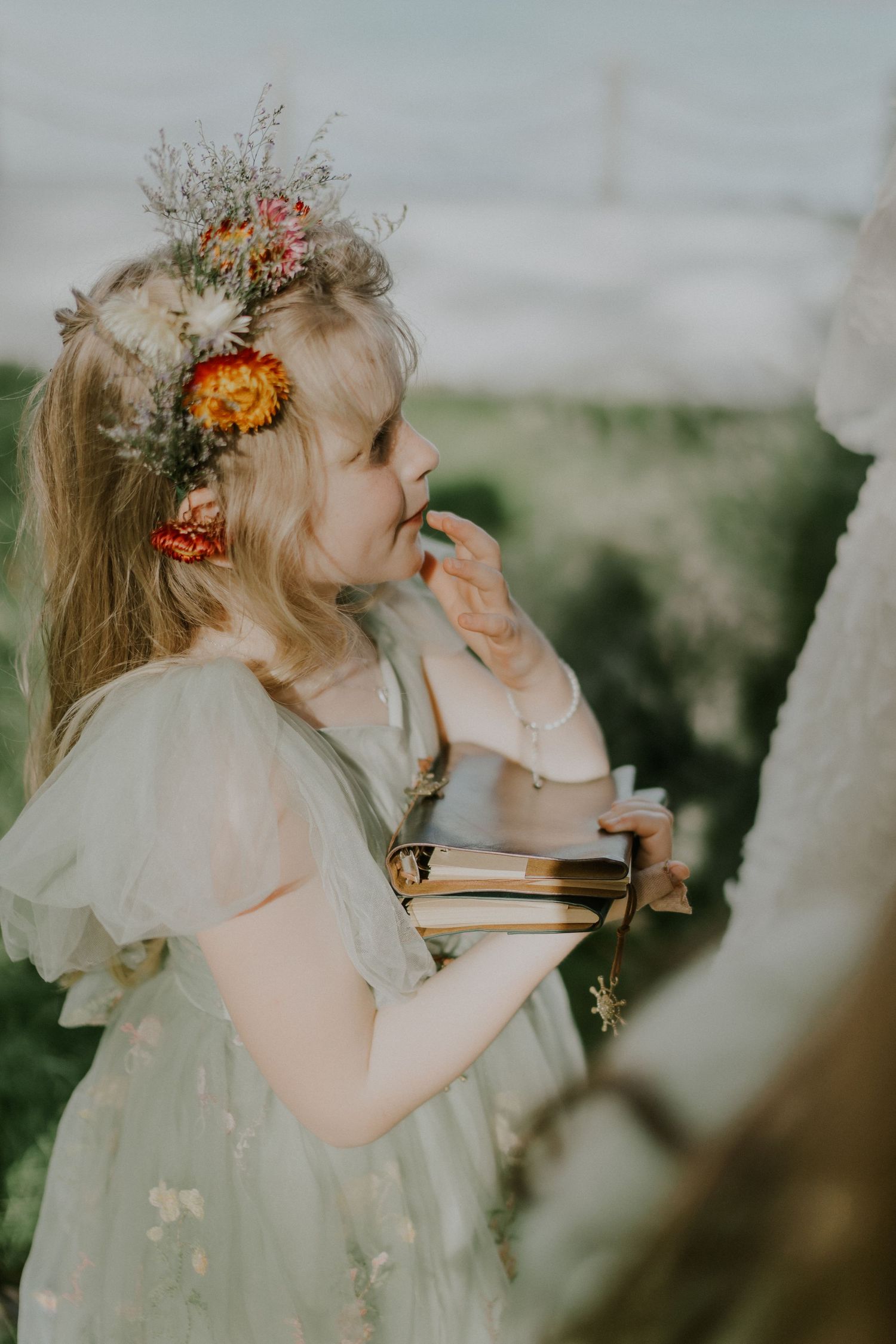 Young flower girl in white dress with floral crown looking up thoughtfully.