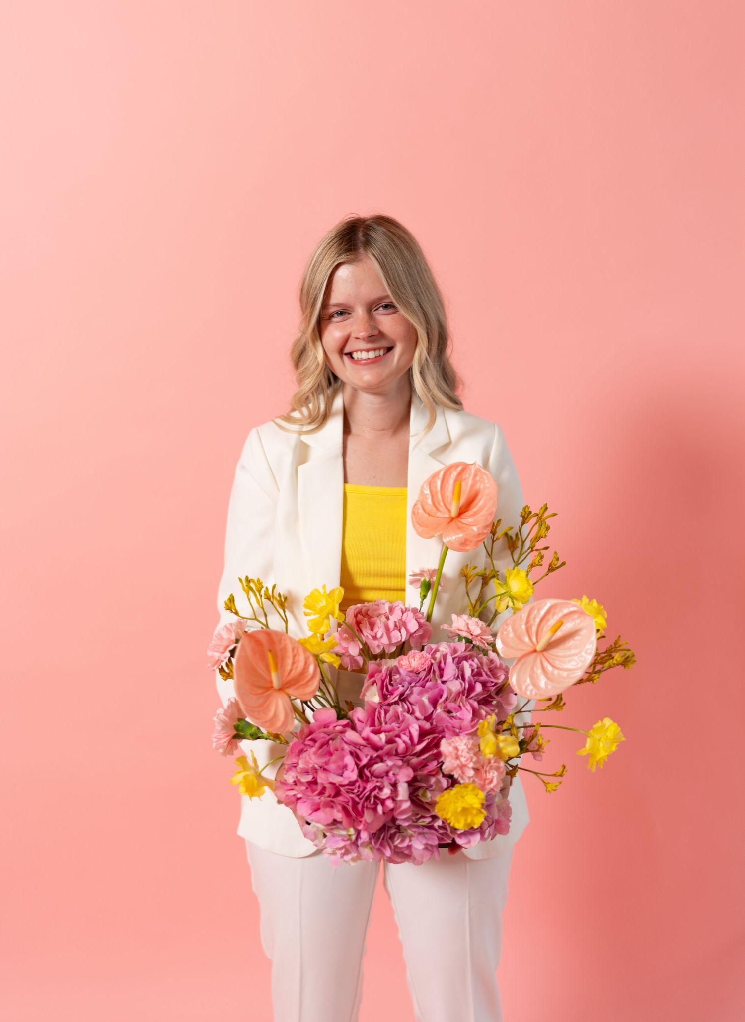 Cheerful person in white suit and yellow top holding vibrant flower arrangement with pink hydrangeas and orange anthuriums.