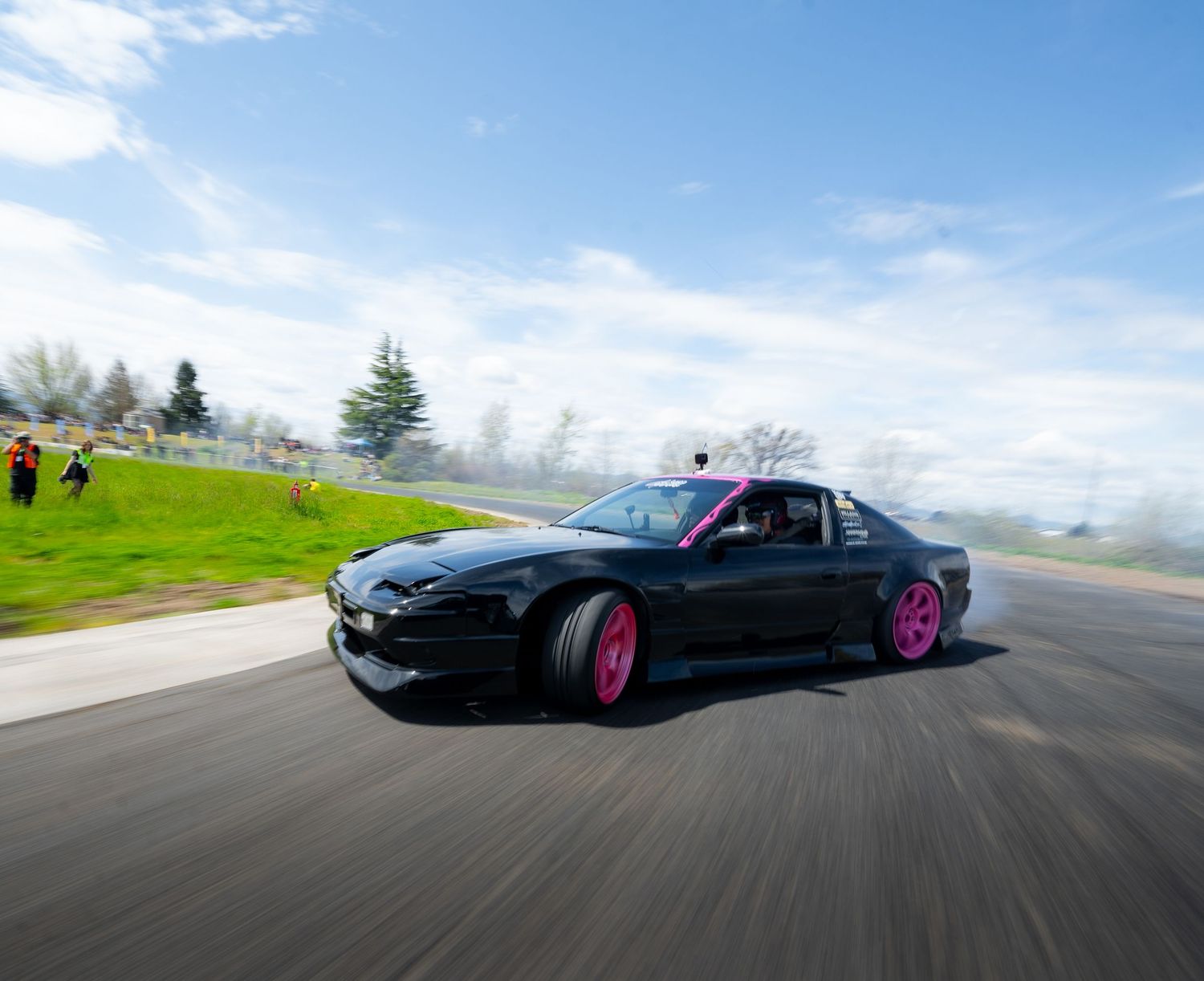 Black sports car with pink wheels drifts around a racetrack curve with mountains in the background.