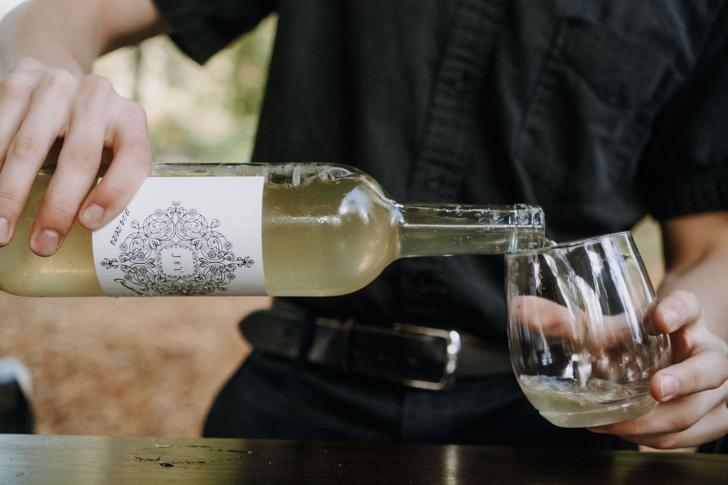 A person pours white wine from an ornately labeled bottle into a stemmed glass outdoors.