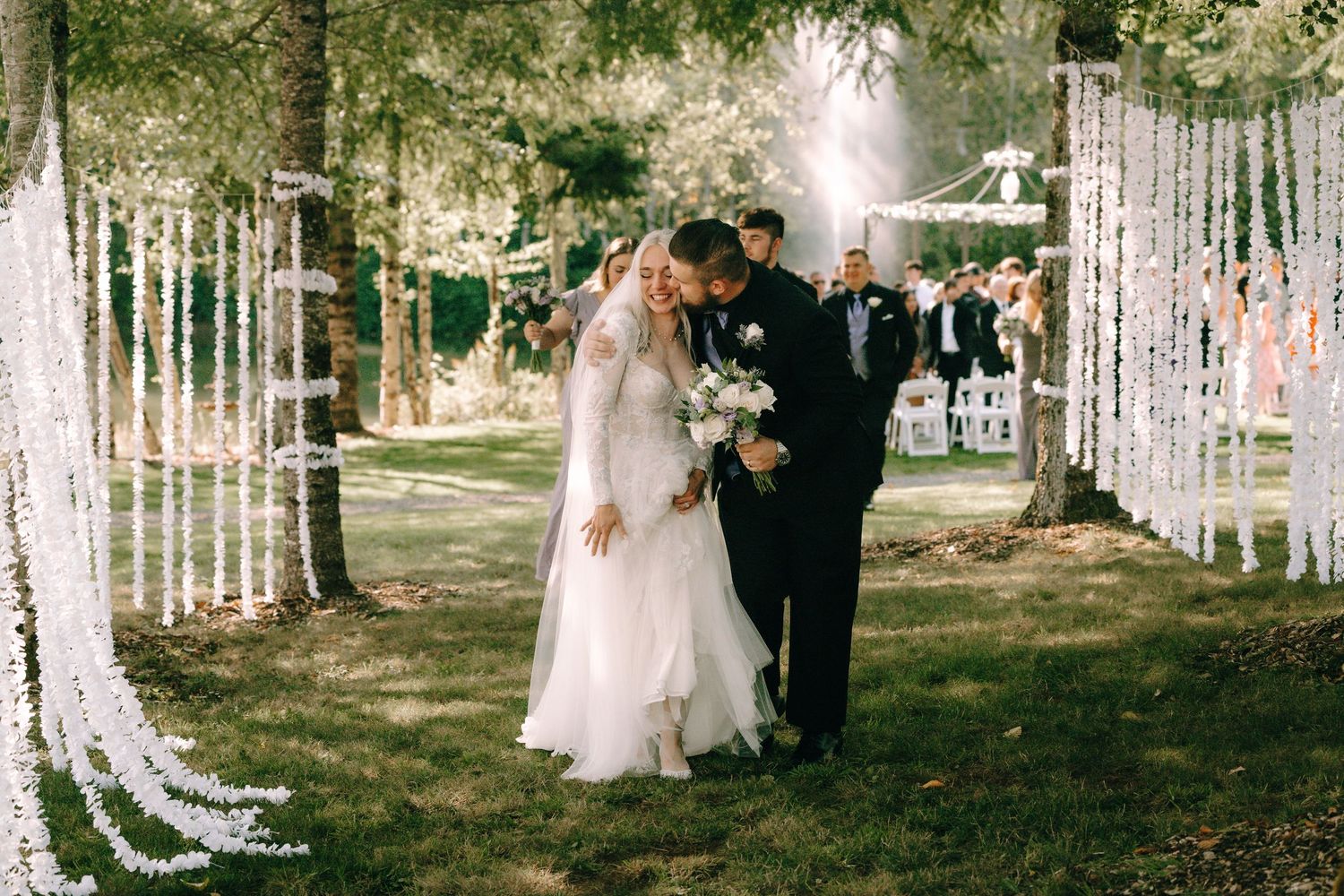 Outdoor wedding ceremony space decorated with hanging white floral arrangements and string lights among trees.