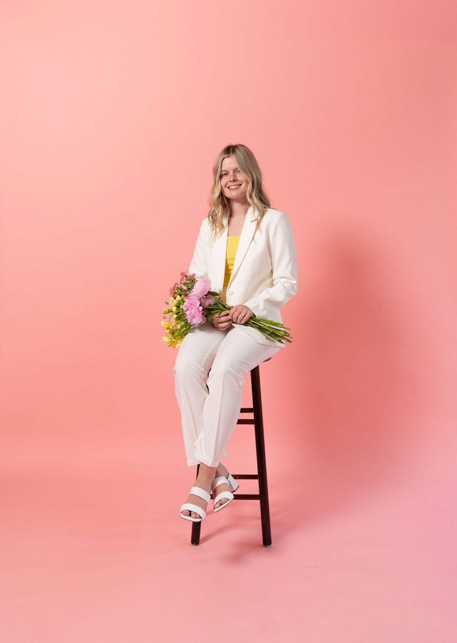 Floral stylist in crisp white suit sits on wooden stool against pink backdrop with fresh flowers.