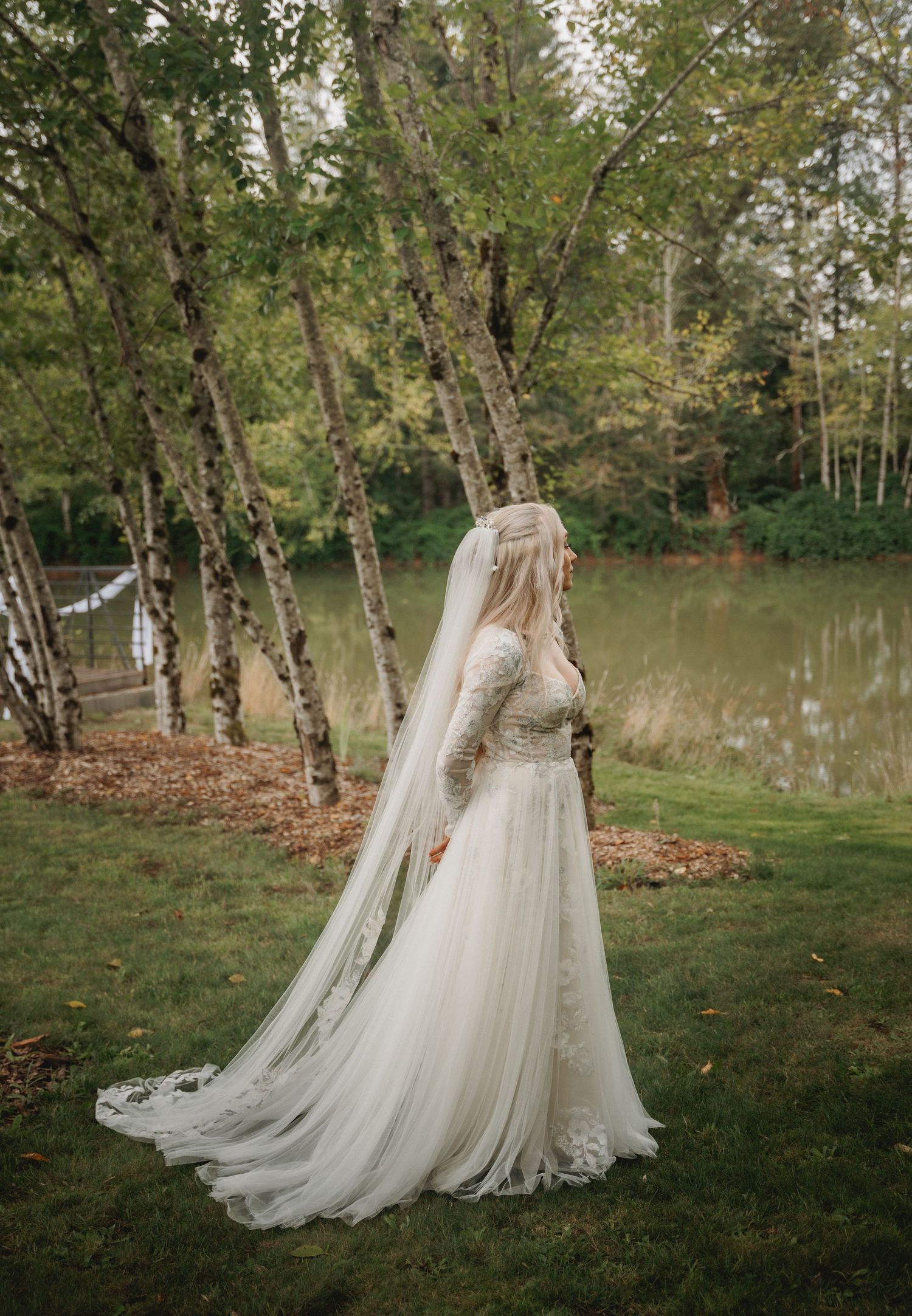 A flowing white wedding dress with long train photographed in a natural outdoor setting with trees and a pond.