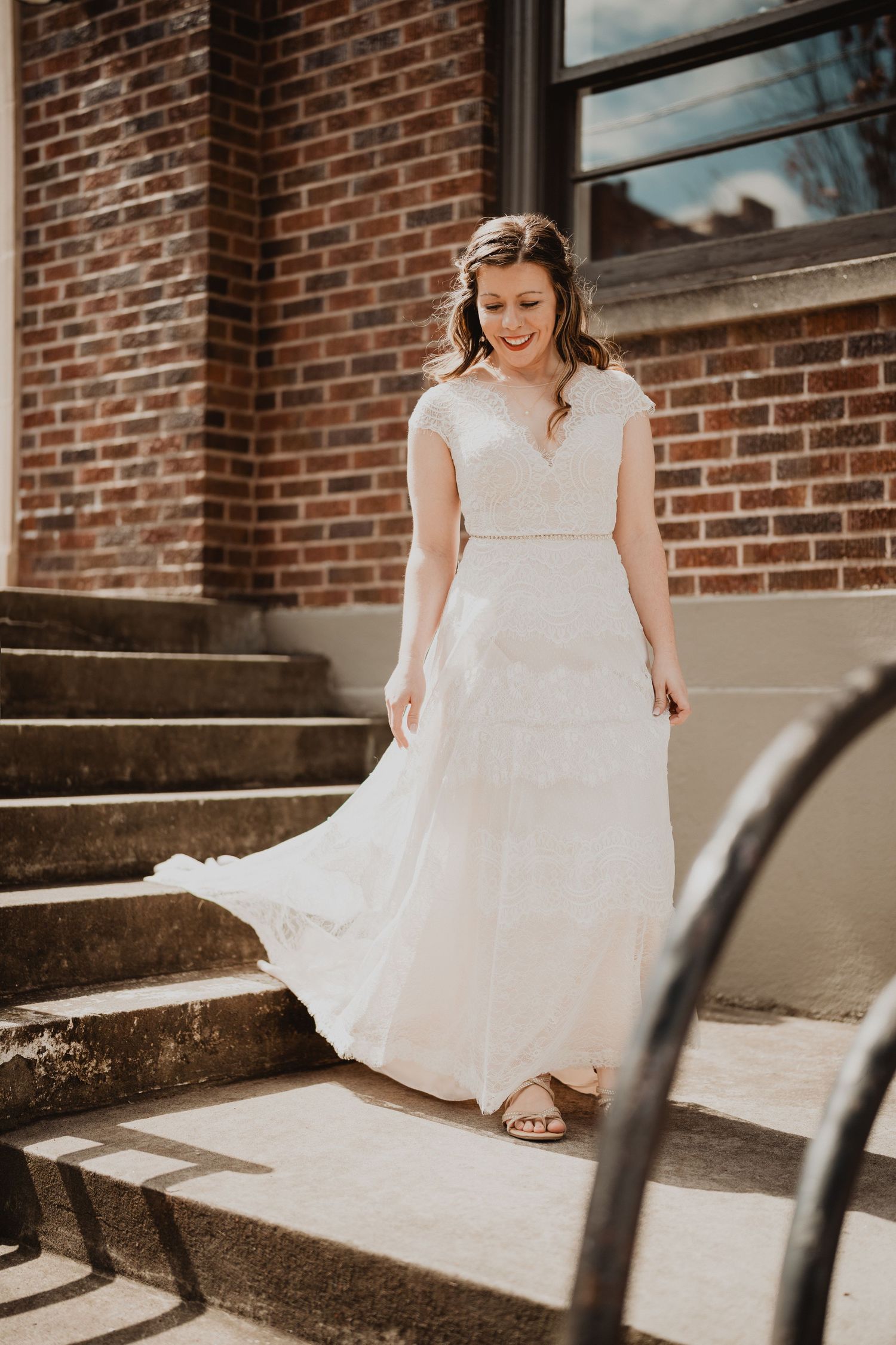 A bride in a flowing white wedding dress walks down sunny concrete steps against a brick building backdrop.