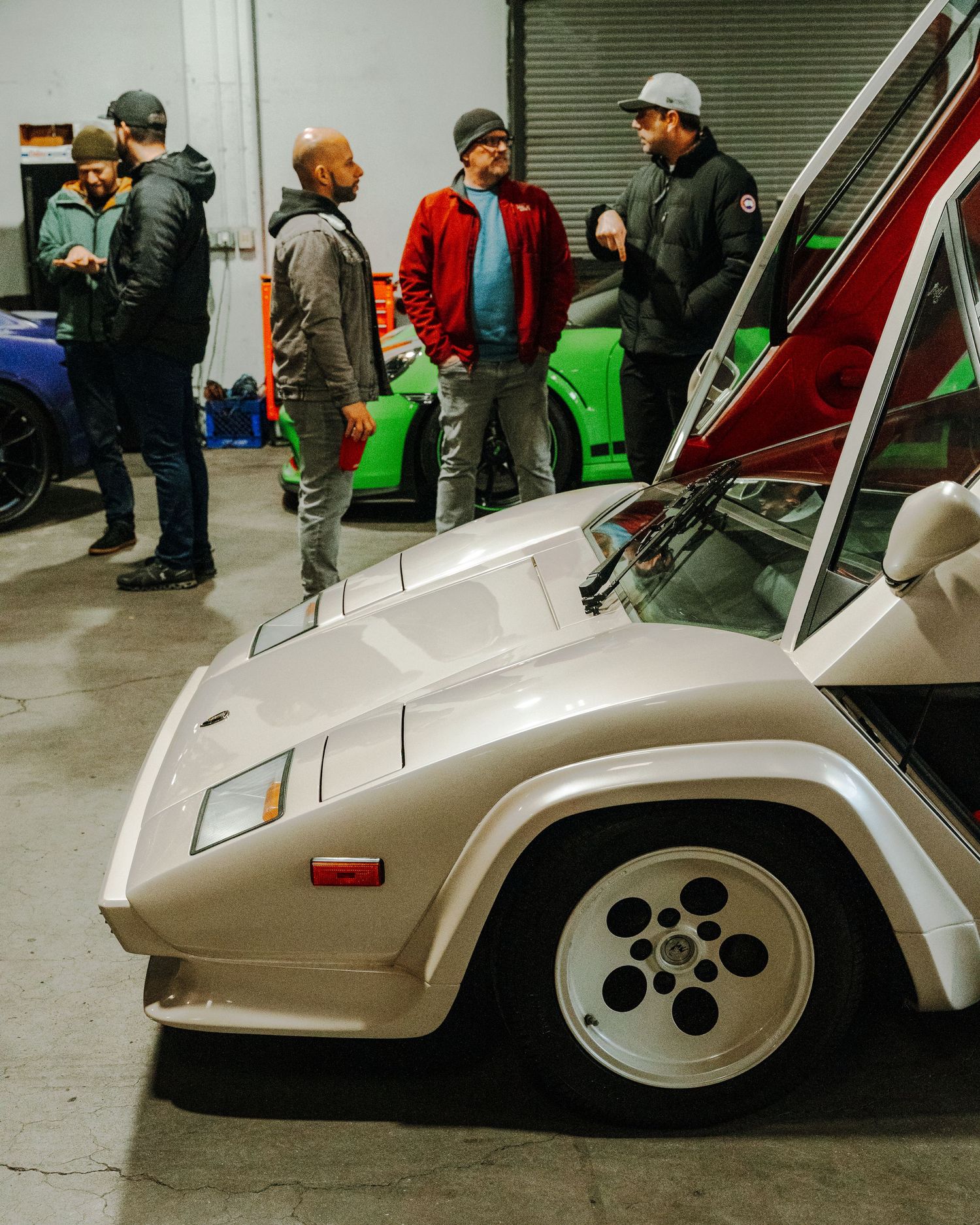 Car enthusiasts gather around a vintage Lamborghini Countach with distinctive silver paint and iconic wheel design in a garage.