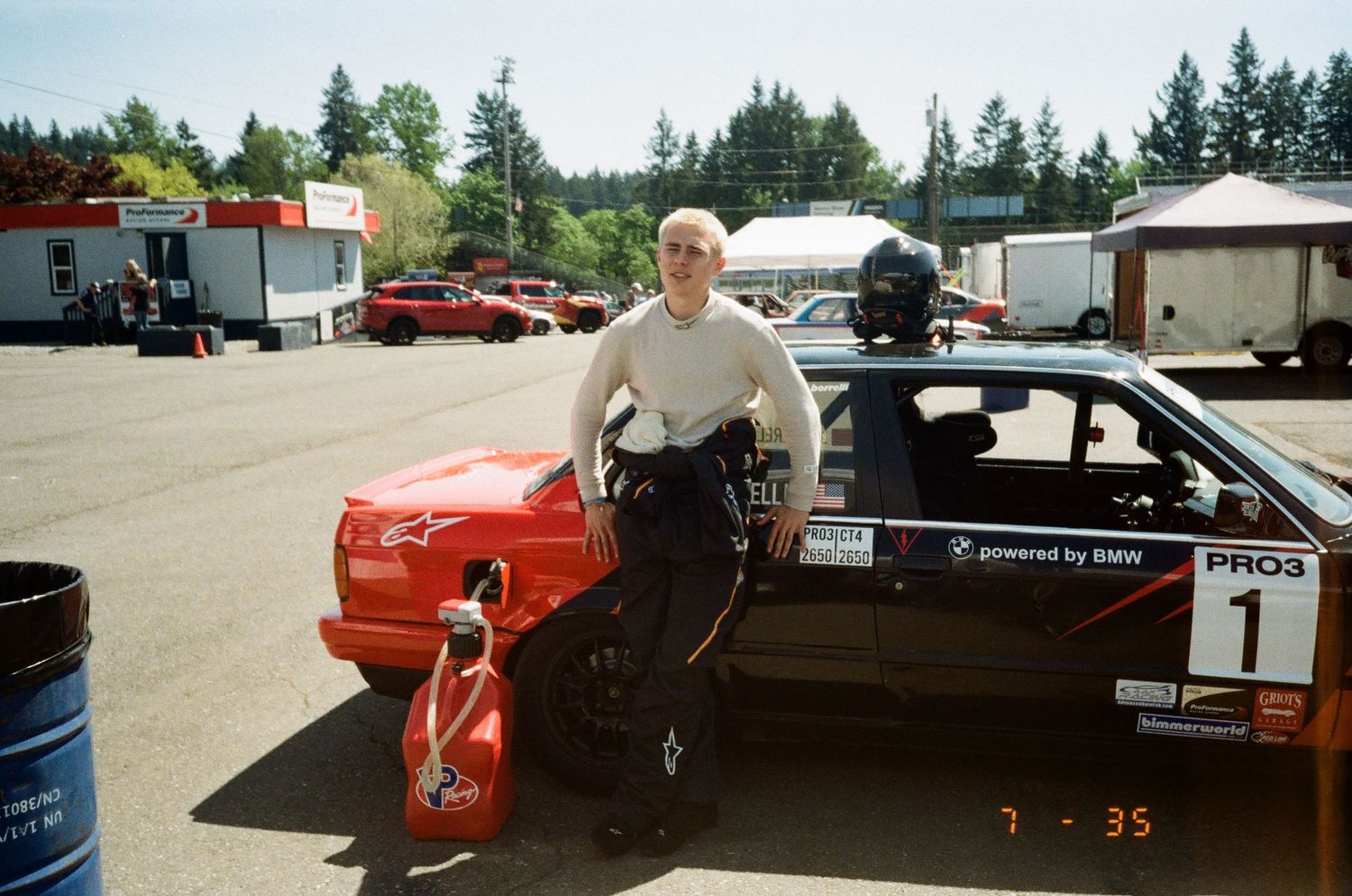 A vintage racing car scene in a parking lot with automotive equipment and trailers visible in the background.