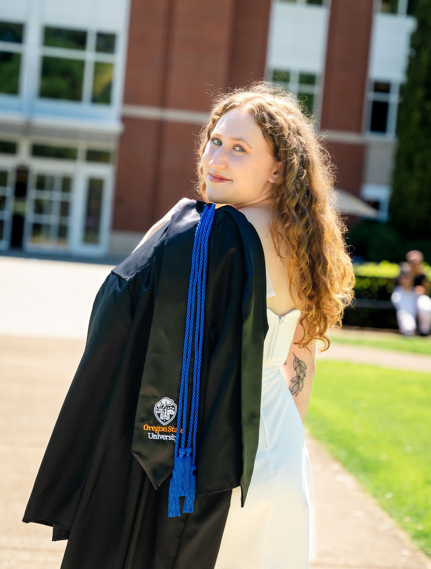 A graduate in academic regalia poses with a black graduation gown and blue stole on a sunny day outside a brick building.