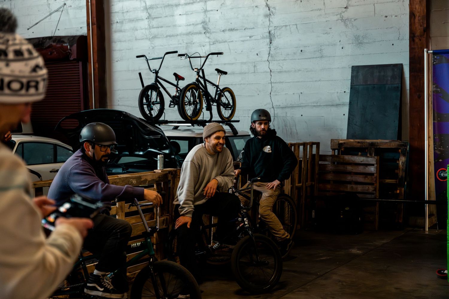 Cyclists gather inside a dimly lit warehouse with bikes silhouetted against a white wall in the background.