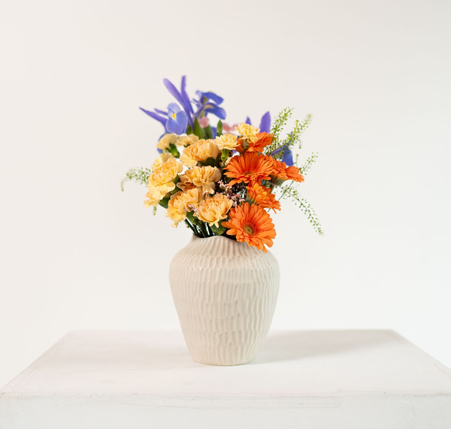 Orange gerbera daisies and purple iris arranged in textured white ceramic vase.