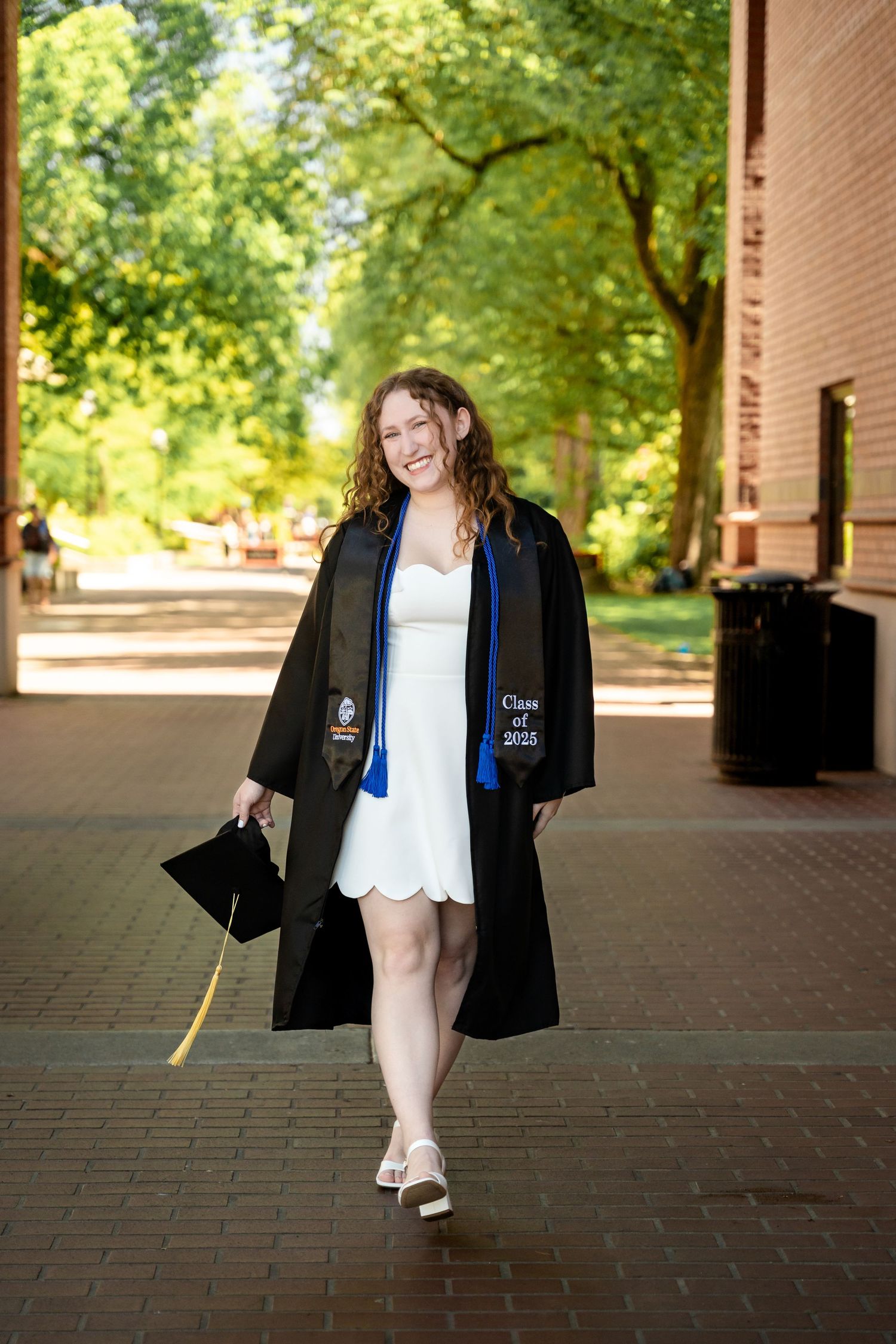 A person in graduation attire walks along a covered brick walkway on a sunny day, surrounded by greenery.