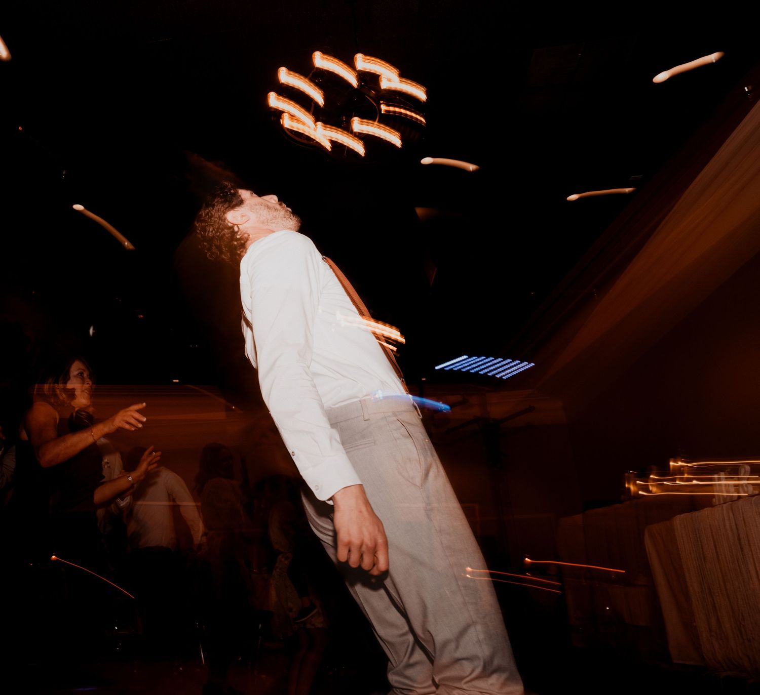 A person in suspenders leans back while dancing at a dimly lit reception venue.