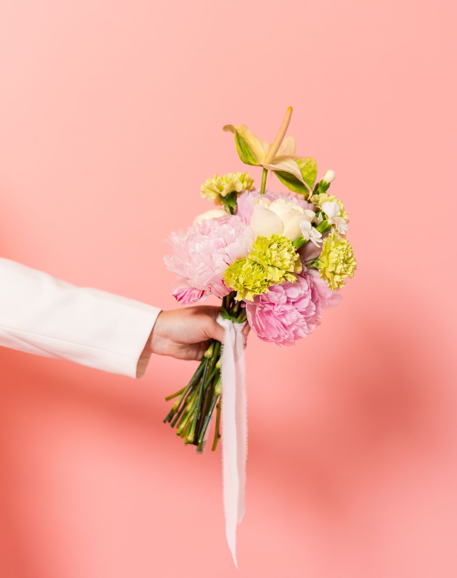 A hand holding a delicate bouquet of pink and green carnations with white ribbon against a coral pink background.
