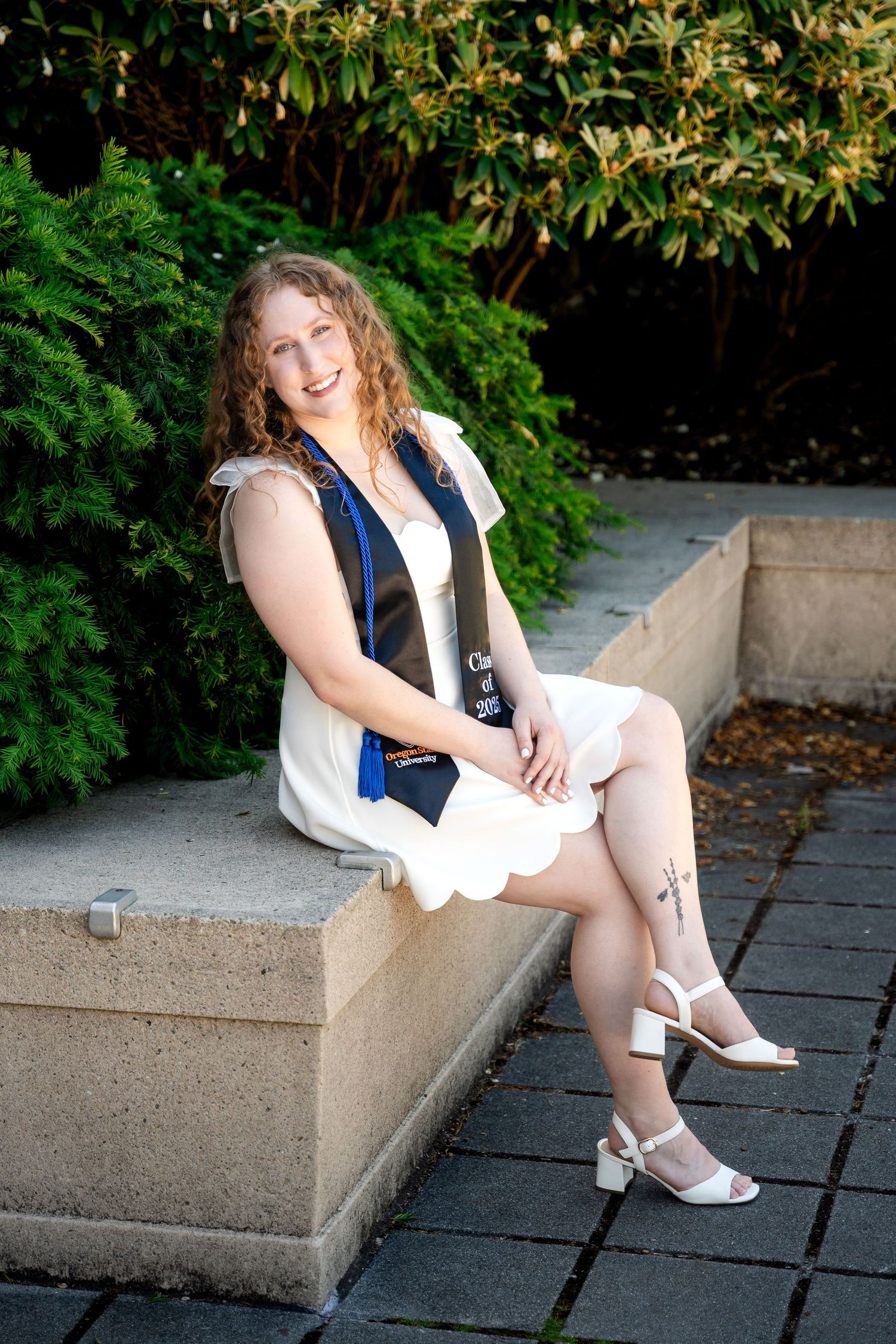 A graduate wearing a navy blue vest and white dress sits on a concrete ledge near greenery and bushes.