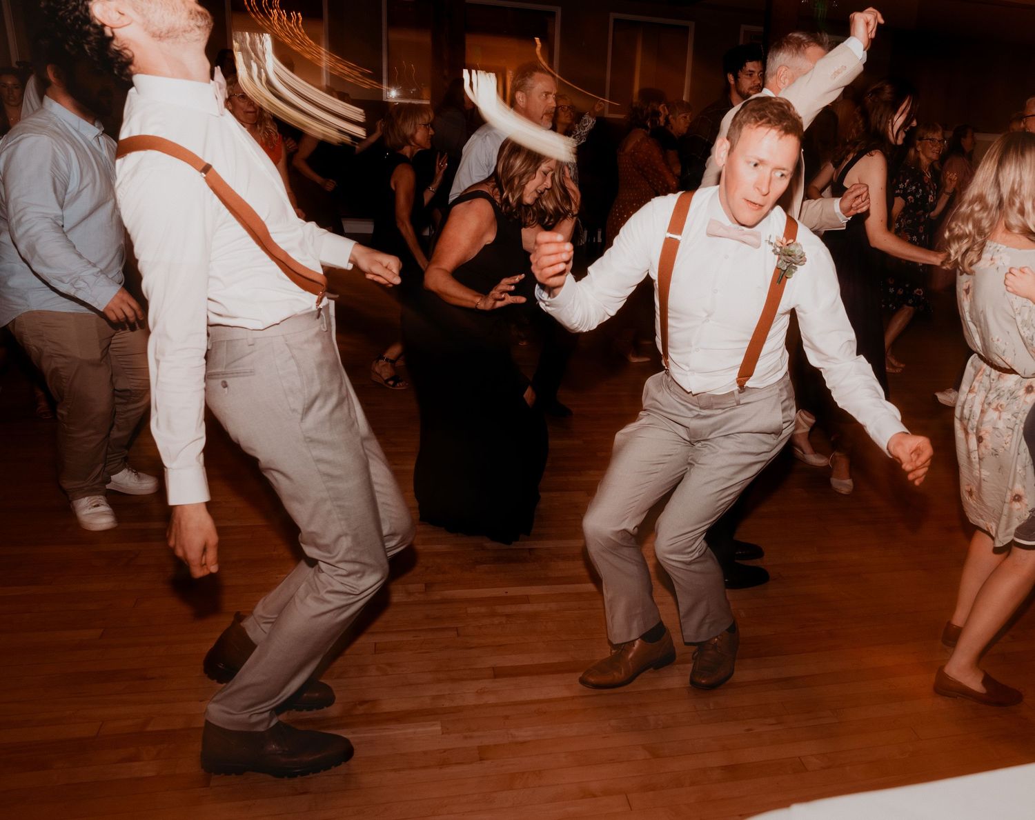 Wedding guests dance energetically on a wooden dance floor at an evening reception celebration.
