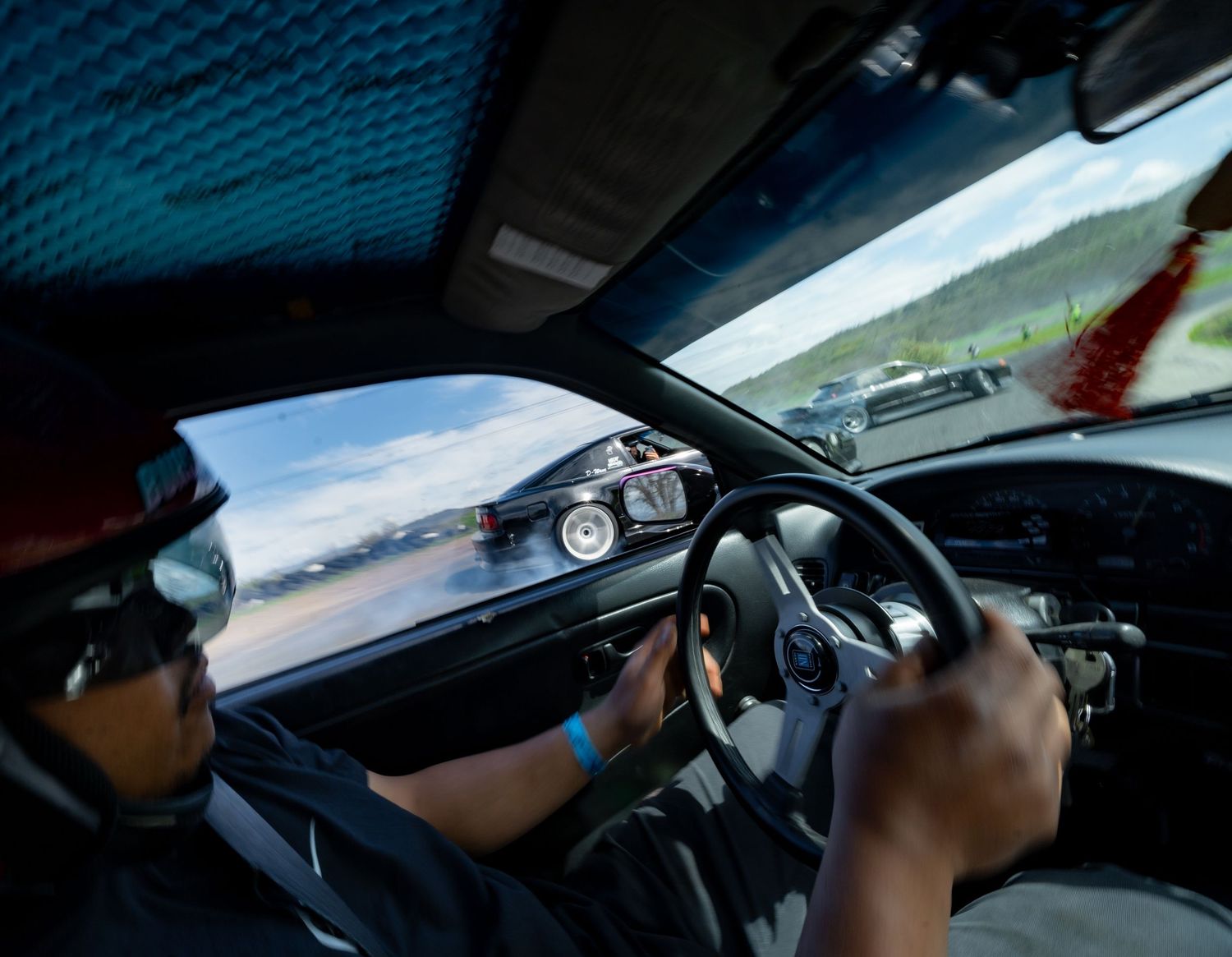 View through interior of car chasing another vehicle on racetrack, showing steering wheel and driver's hands.