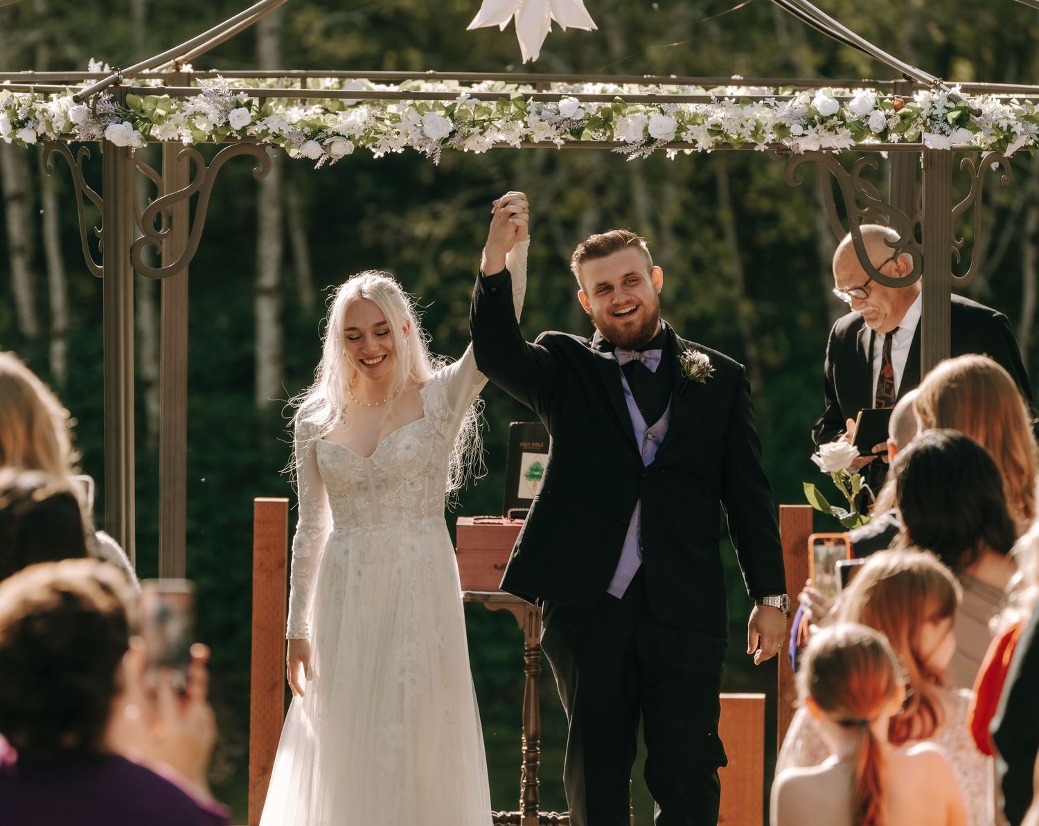 Outdoor wedding ceremony under floral arch decorations with guests seated.
