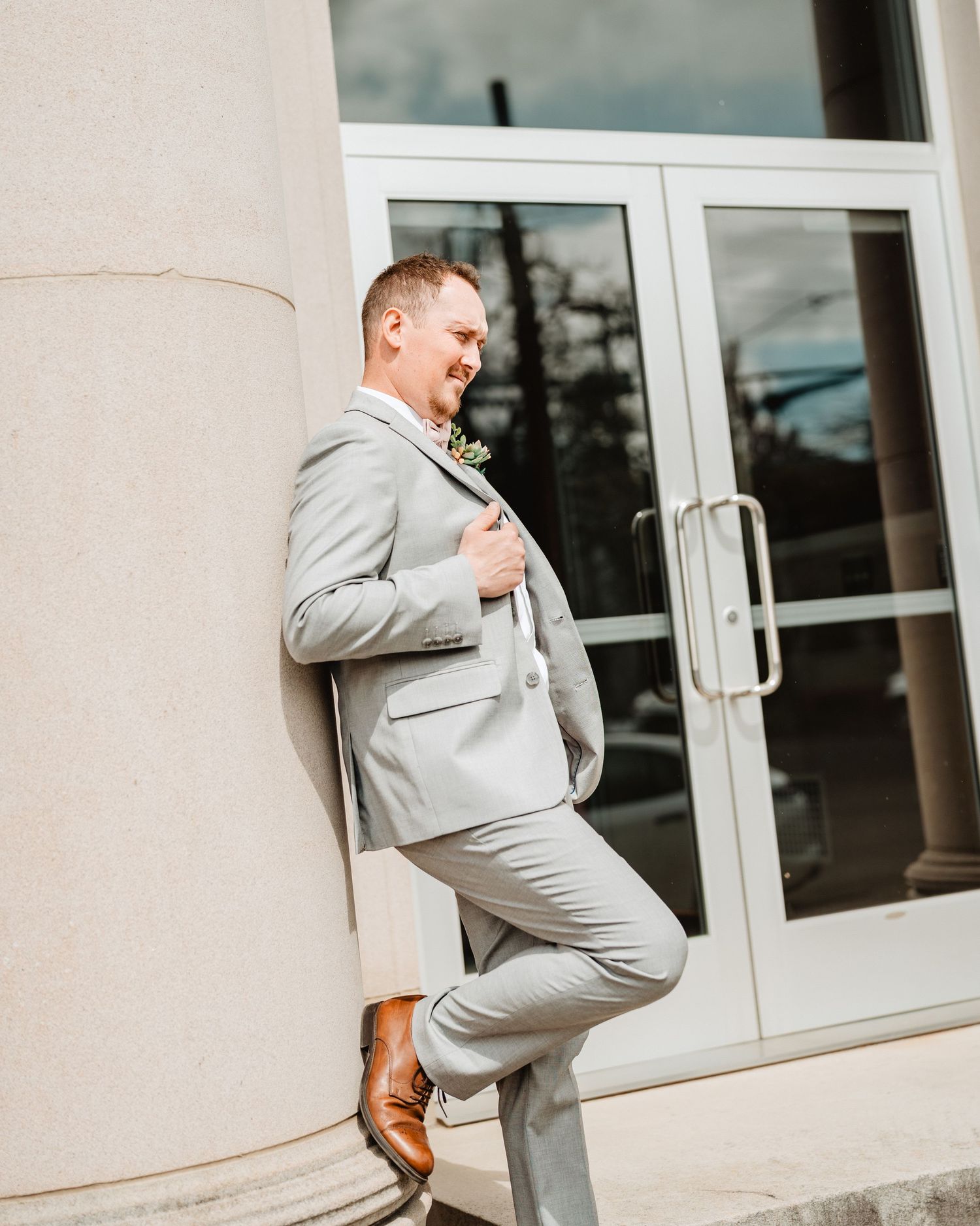 A person wearing a light gray suit strikes an energetic pose while leaning against a building with glass doors.