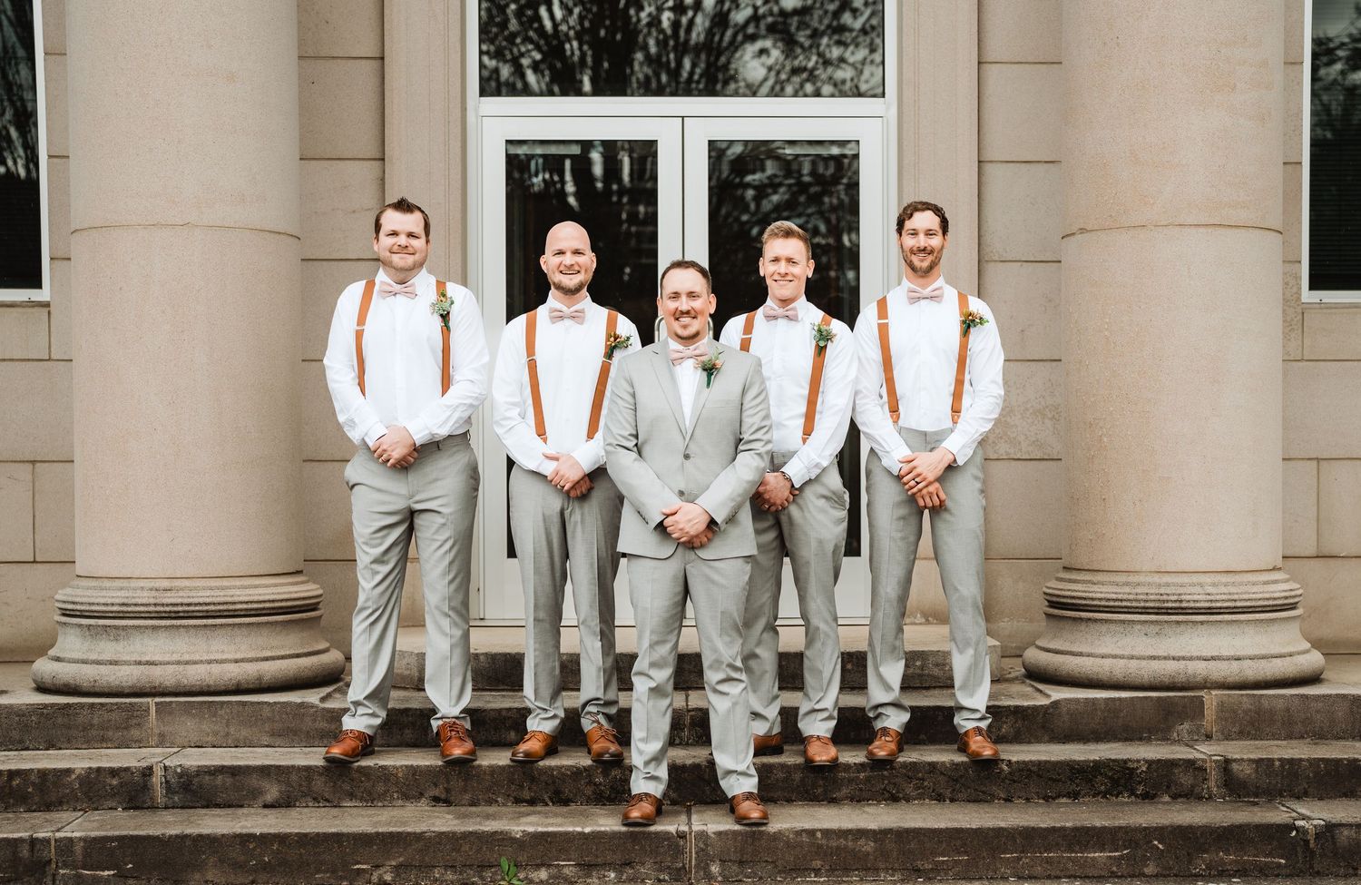 Wedding party in white attire sits casually on a stone staircase between classical columns.