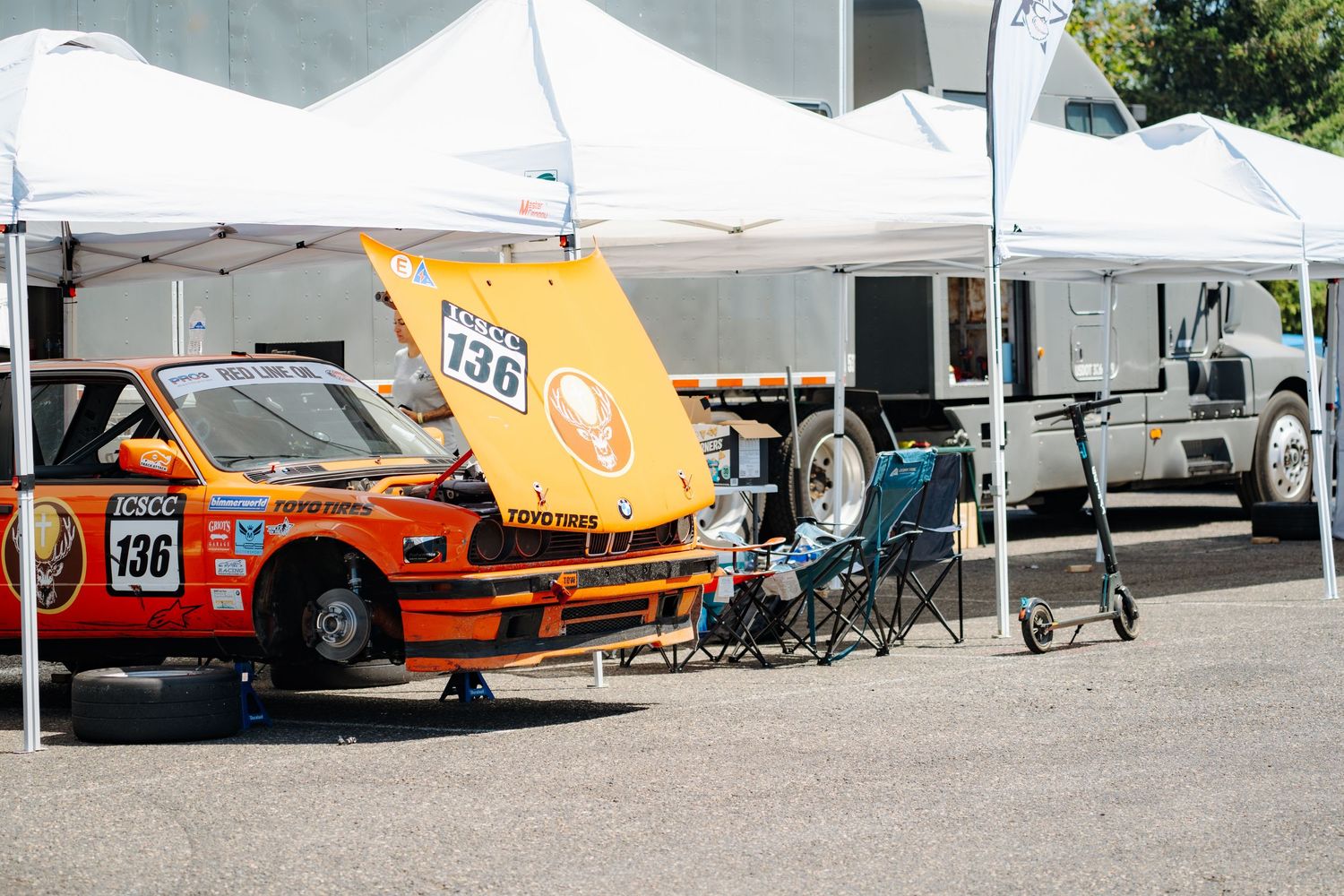 Row of racing cars and trailers parked in a pit lane with orange and white vehicles visible in the lineup.