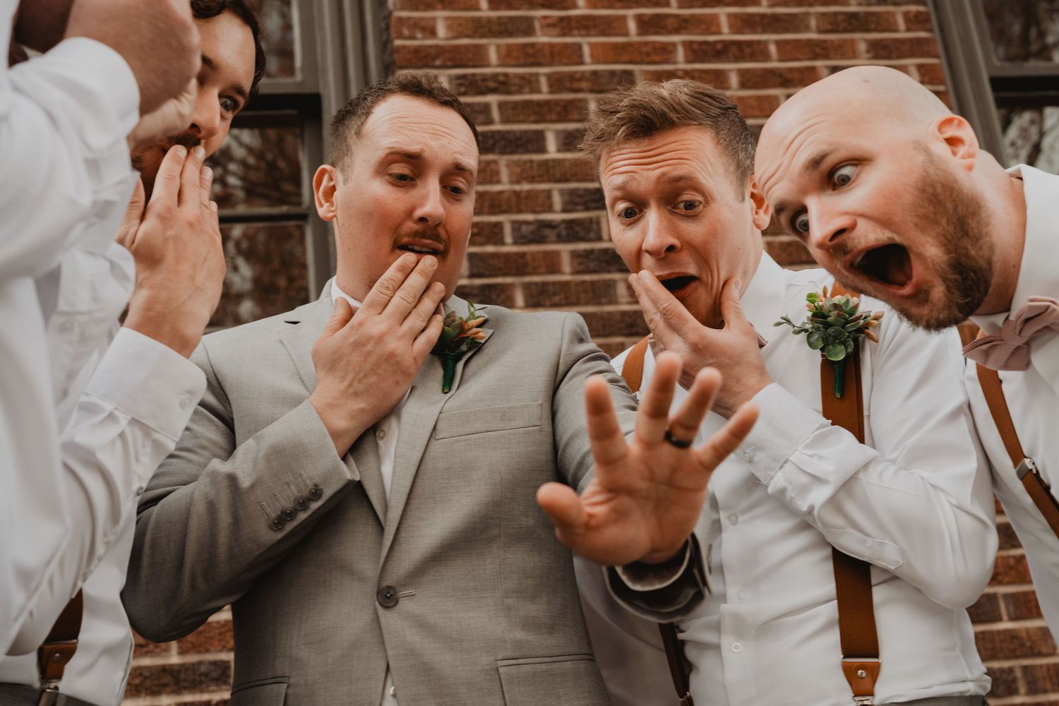 Groomsmen in white shirts and suspenders react with exaggerated expressions of shock as they pose together.