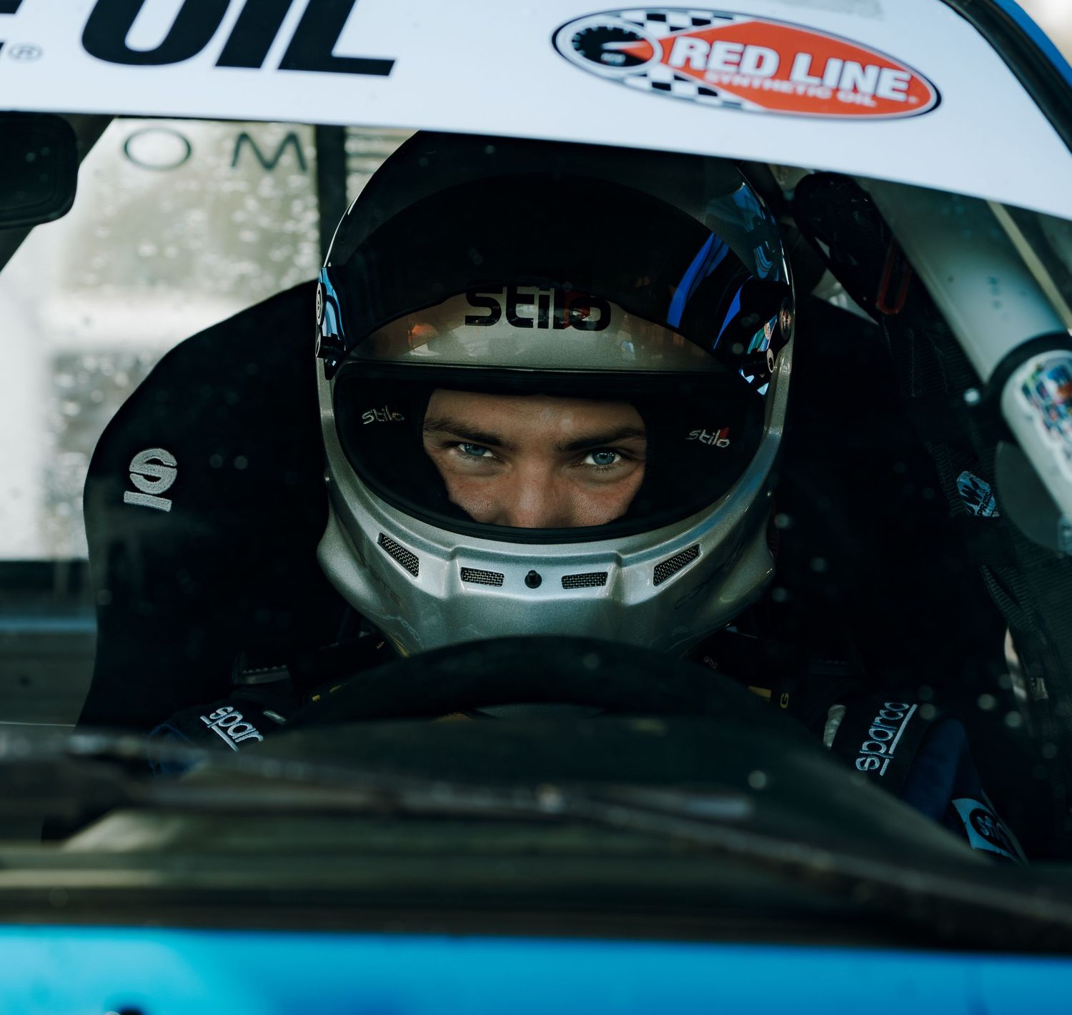 Professional racing driver visible through helmet visor and safety equipment in motorsport vehicle cockpit.