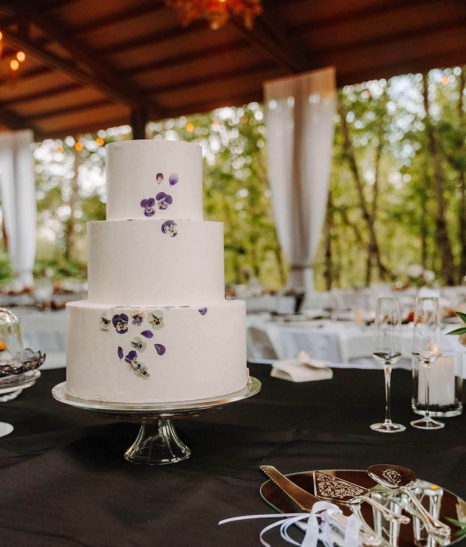 Three-tiered white wedding cake decorated with purple flower petals displayed on a glass stand at a reception venue.