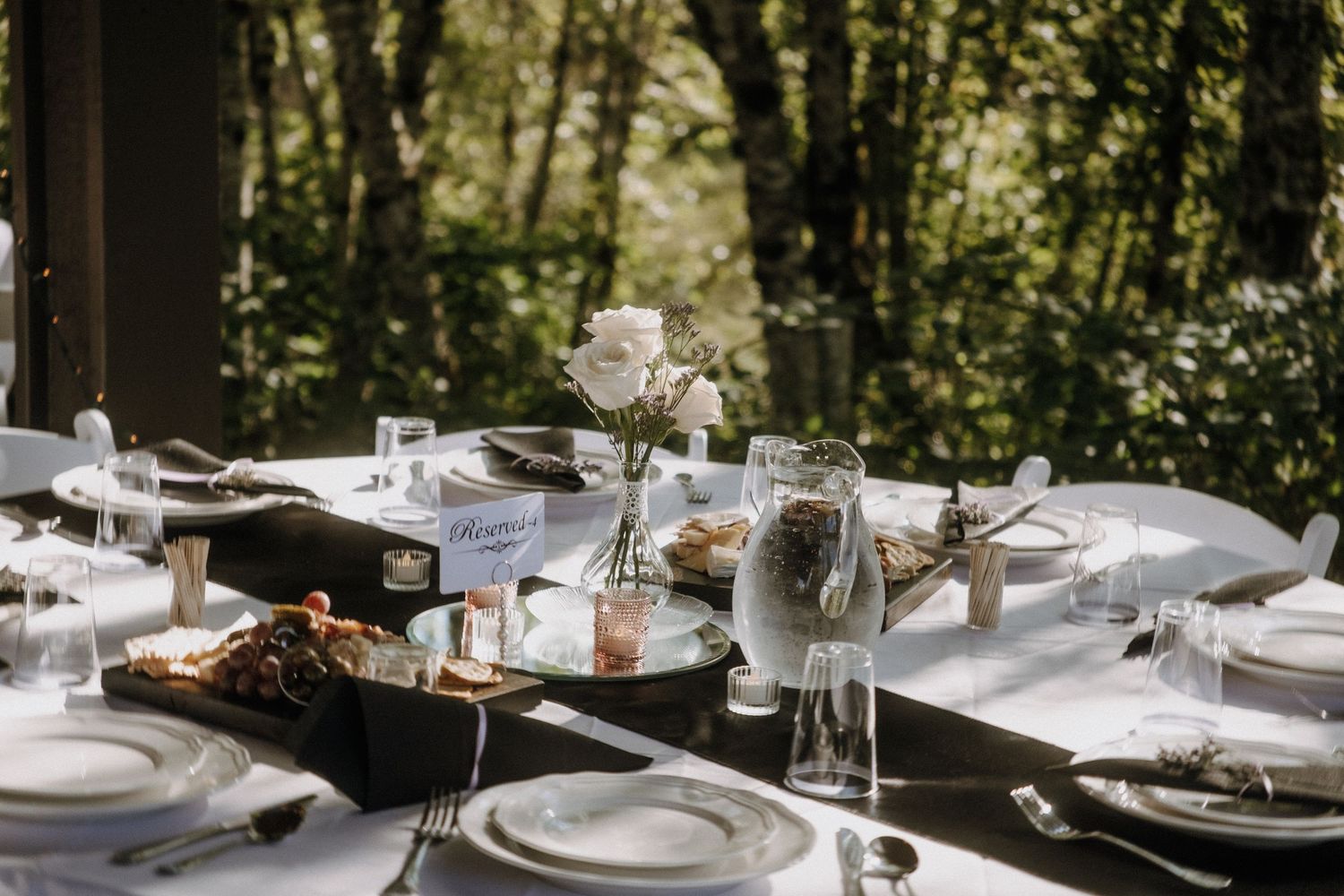 An elegant outdoor dining table set with white plates, glassware, and floral centerpieces in a wooded setting.