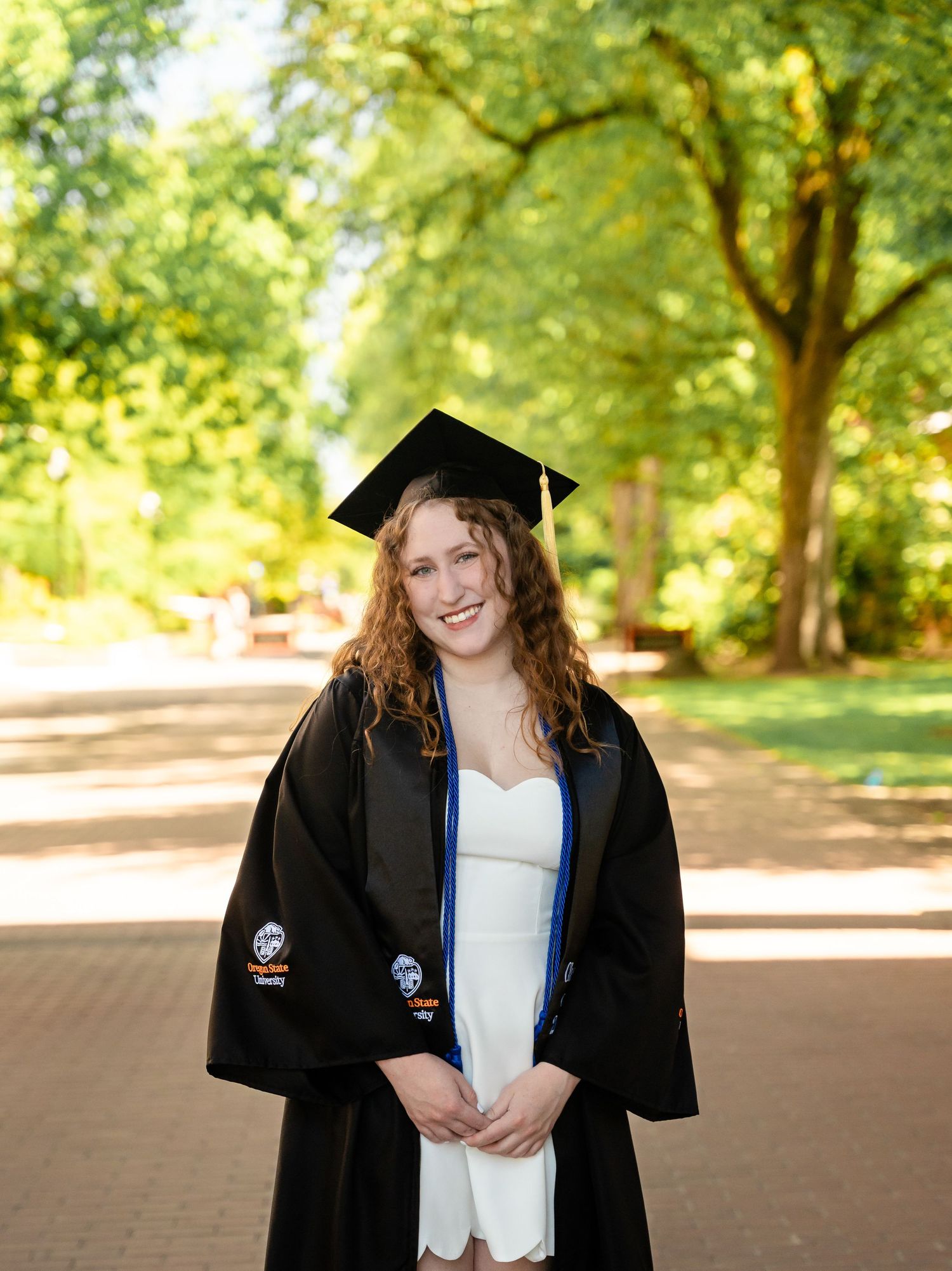 A graduate in a black cap and gown stands smiling on a tree-lined brick pathway during commencement season.