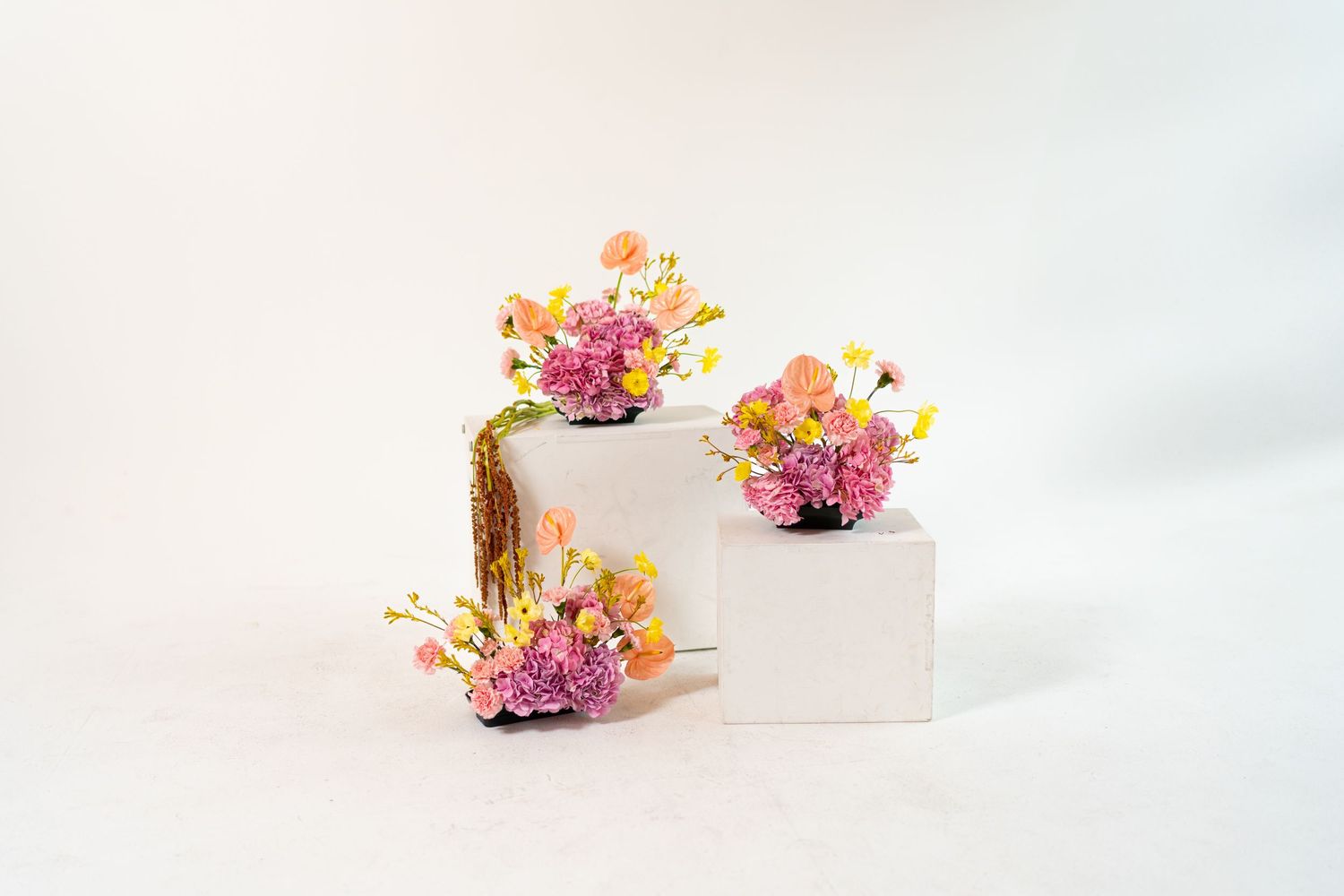 Three colorful flower arrangements displayed on white and wooden pedestals against light background.