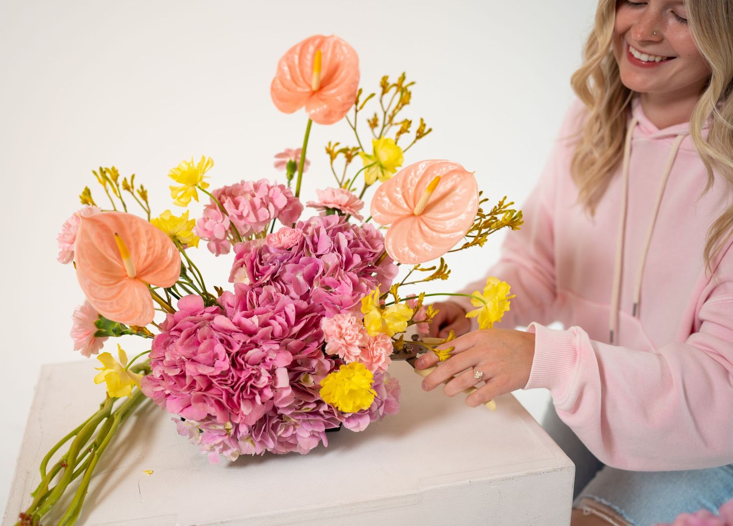 Person in pink shirt arranging colorful flowers including pink hydrangeas and coral-colored blooms on white surface.