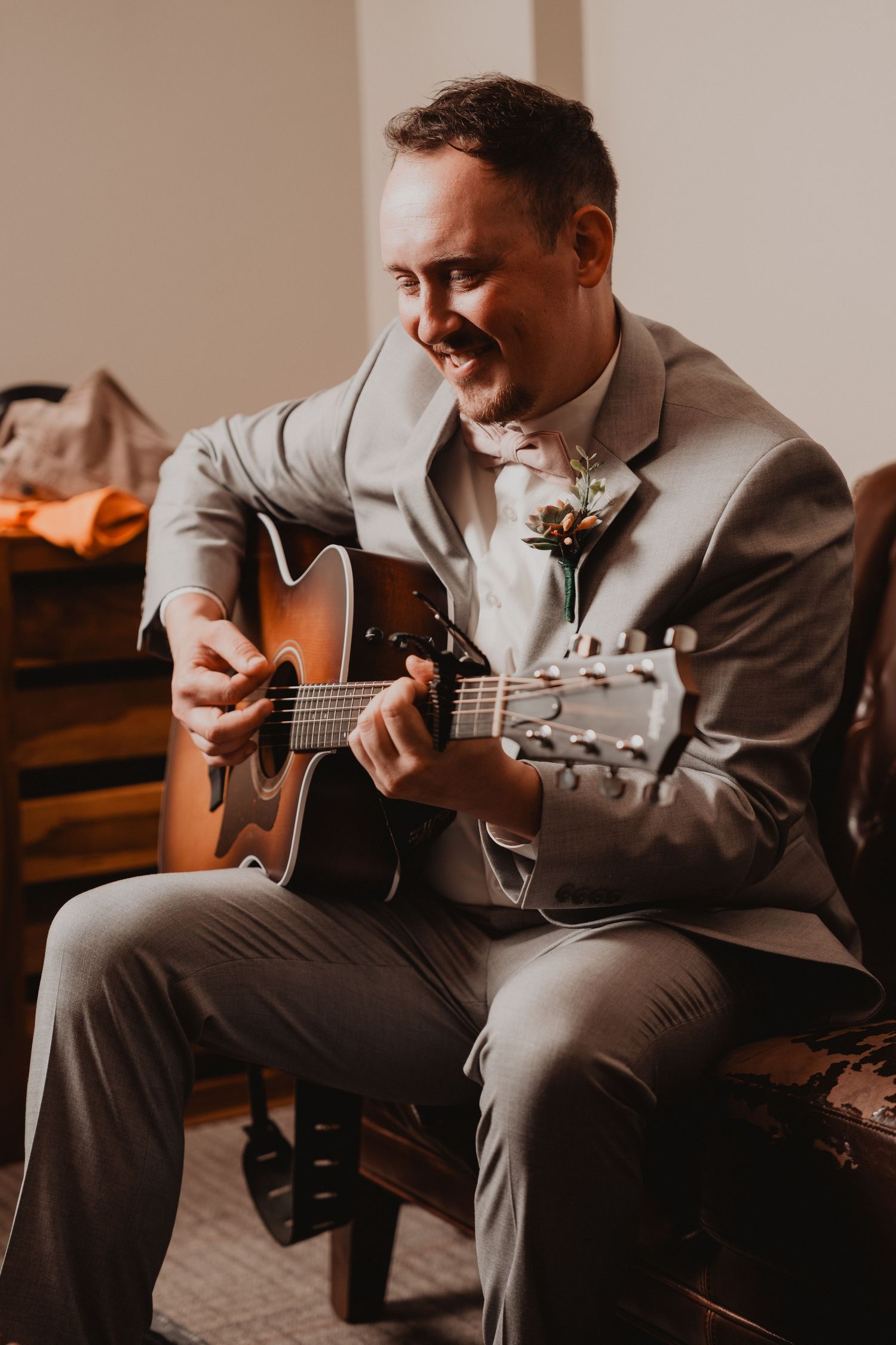 A groom in a light gray suit smiles while playing acoustic guitar during a wedding celebration.