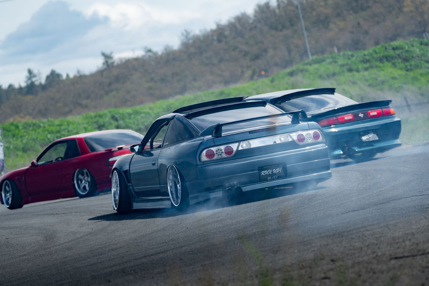 Cars perform synchronized drifting maneuvers in formation on a curved mountain track during an overcast day.