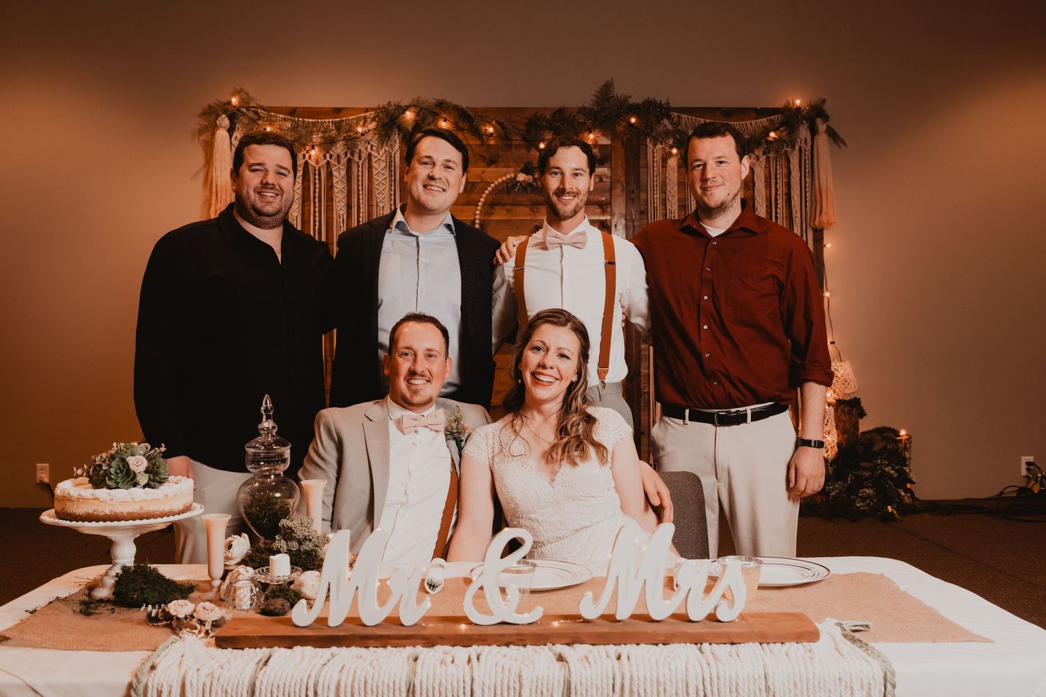 A group in formal attire poses behind a rustic wooden table with decorative 'Mr & Mrs' sign at a wedding reception.