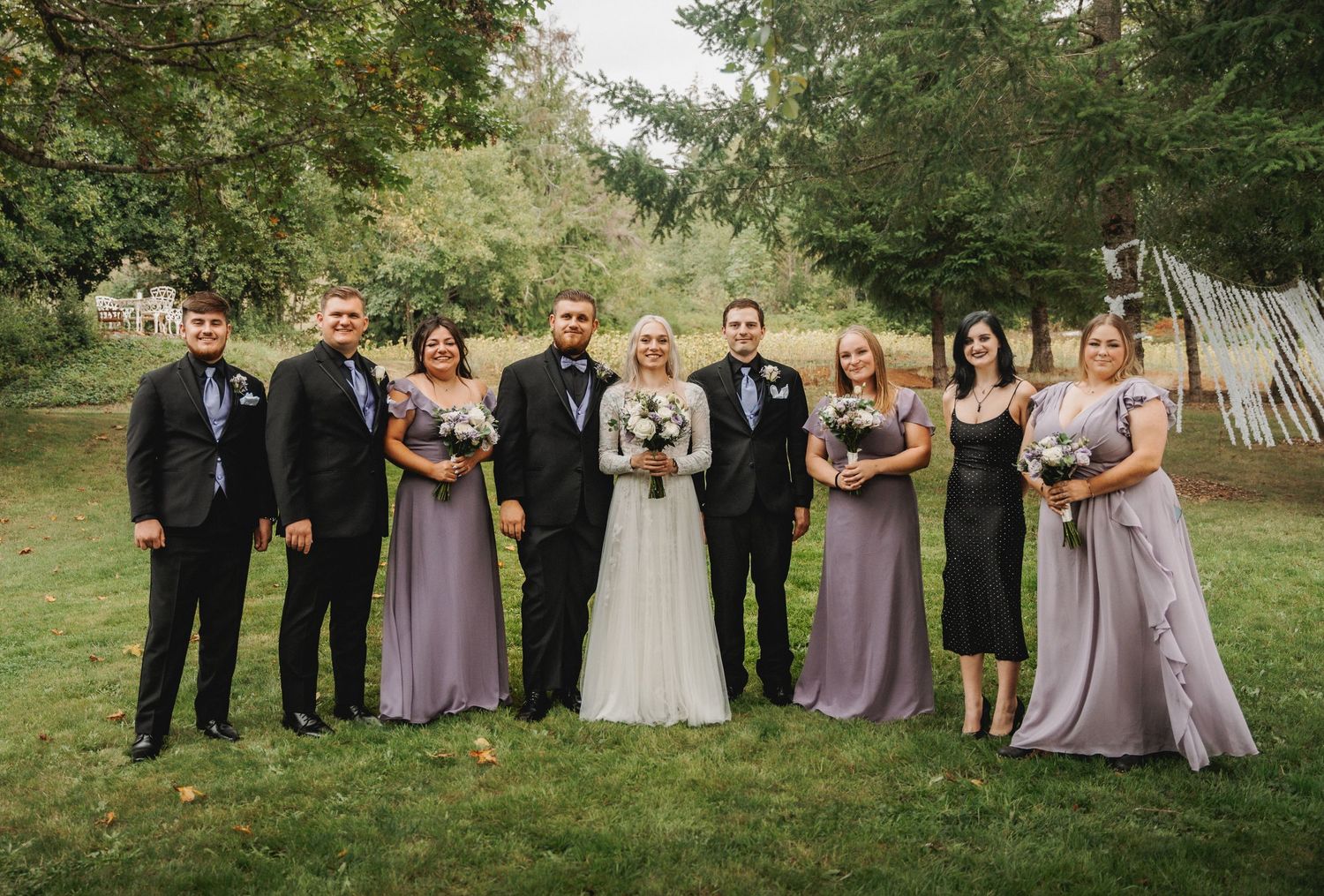 Wedding party in purple and black attire poses together on grassy lawn.