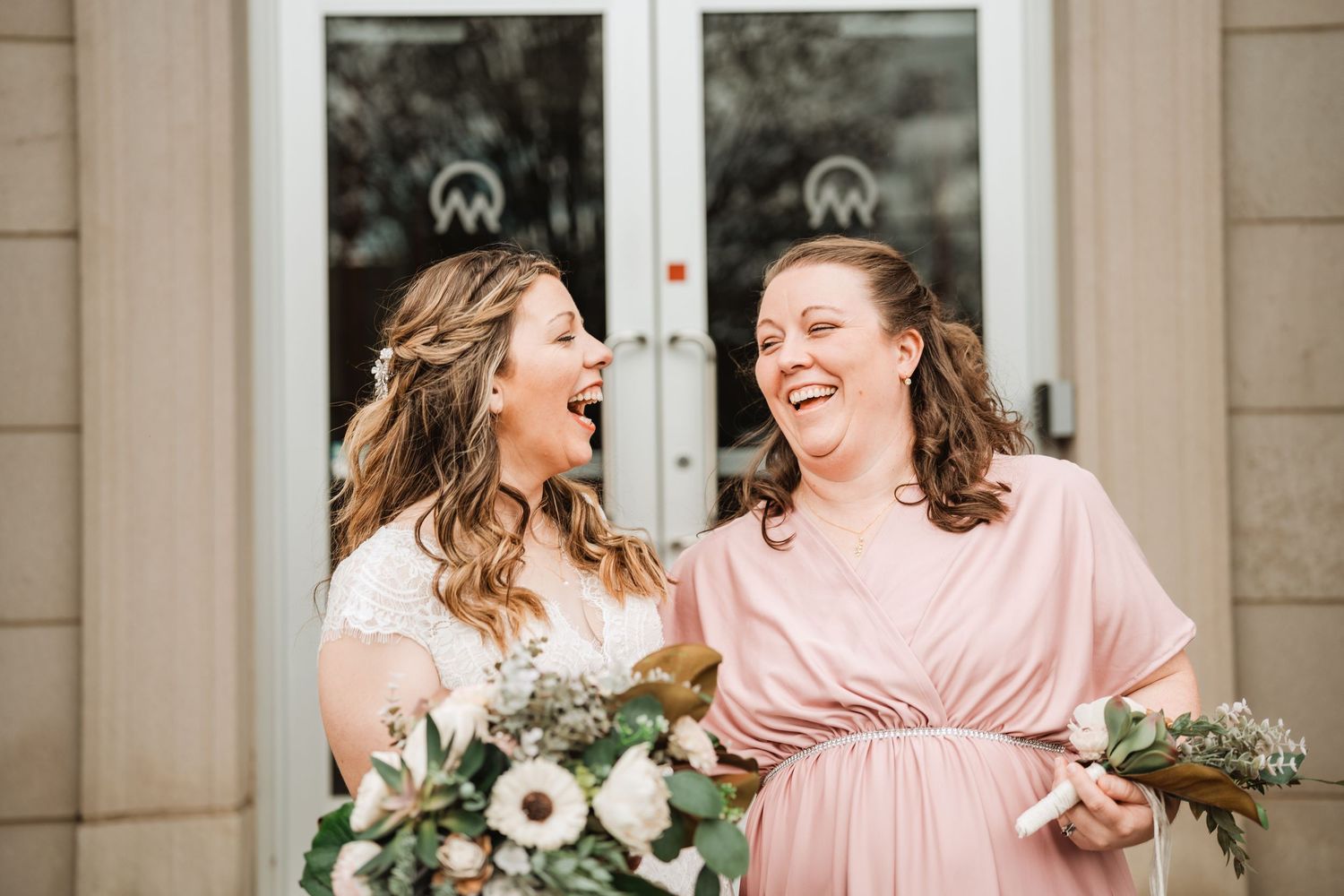 Bridesmaids in blush pink dresses share a candid laughing moment together at an outdoor venue.