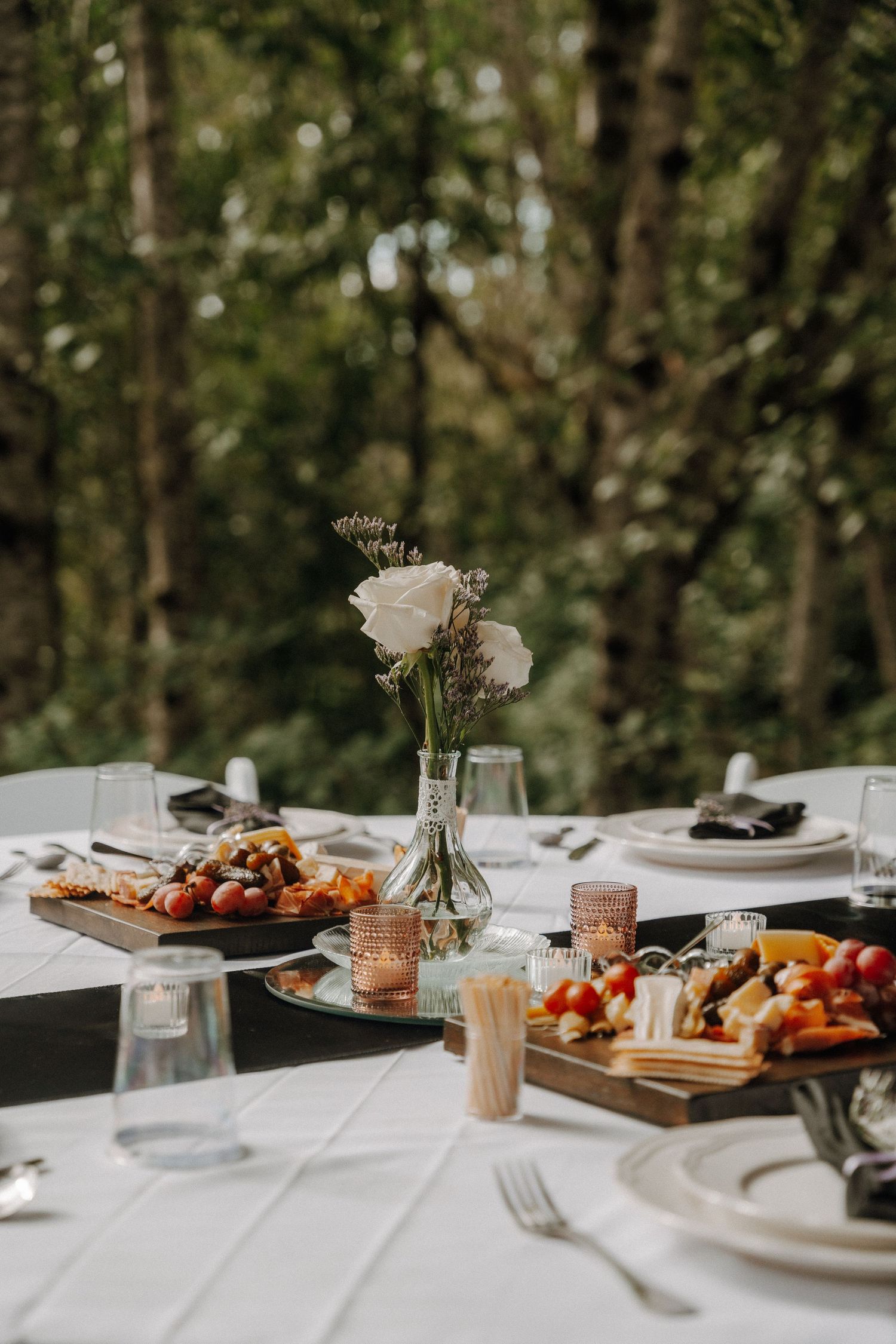Elegant wedding table setting with white roses in vase, plates of appetizers, and rustic outdoor decor in forest setting.