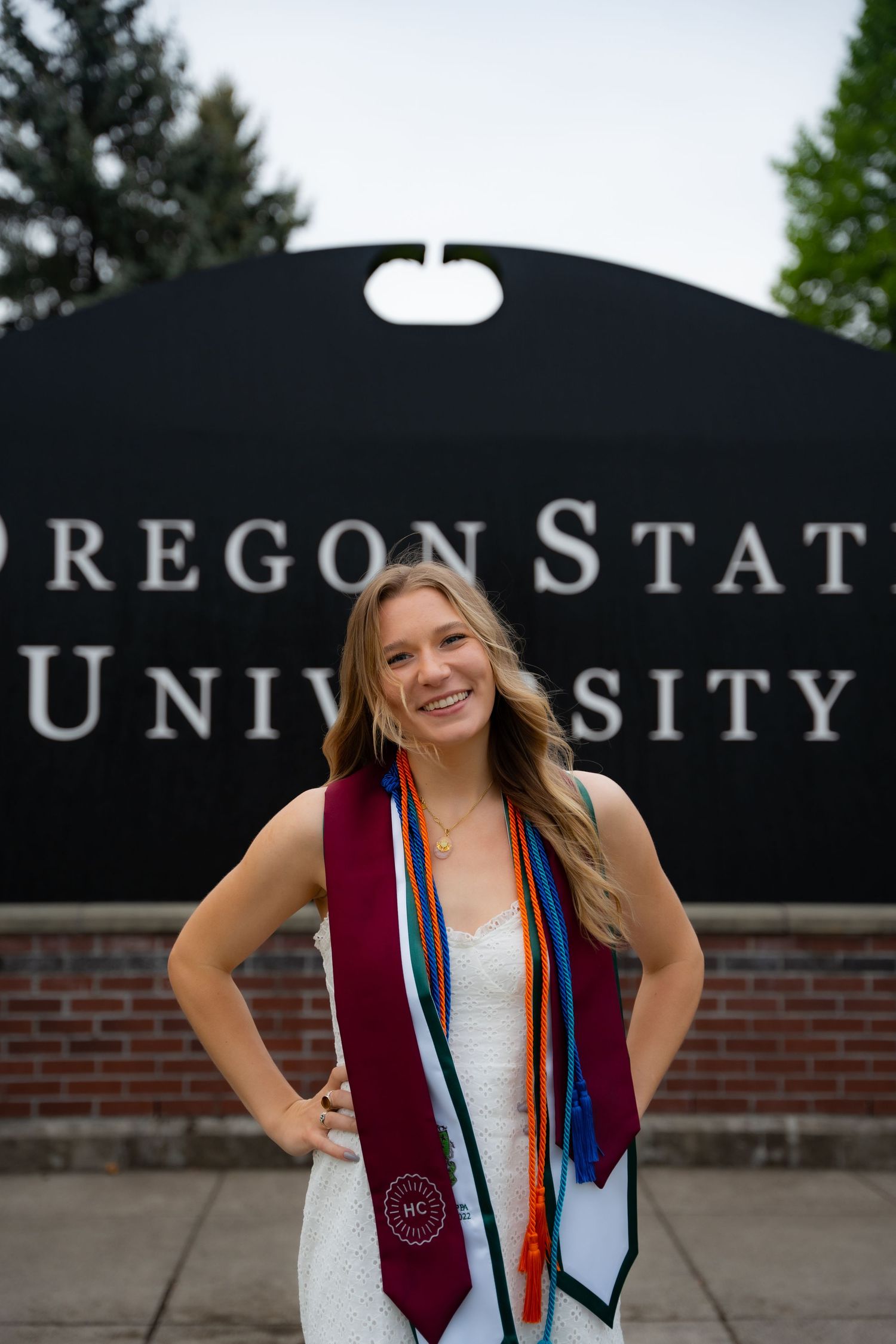 Graduate wearing decorative stoles poses in front of Oregon State University entrance sign on graduation day.