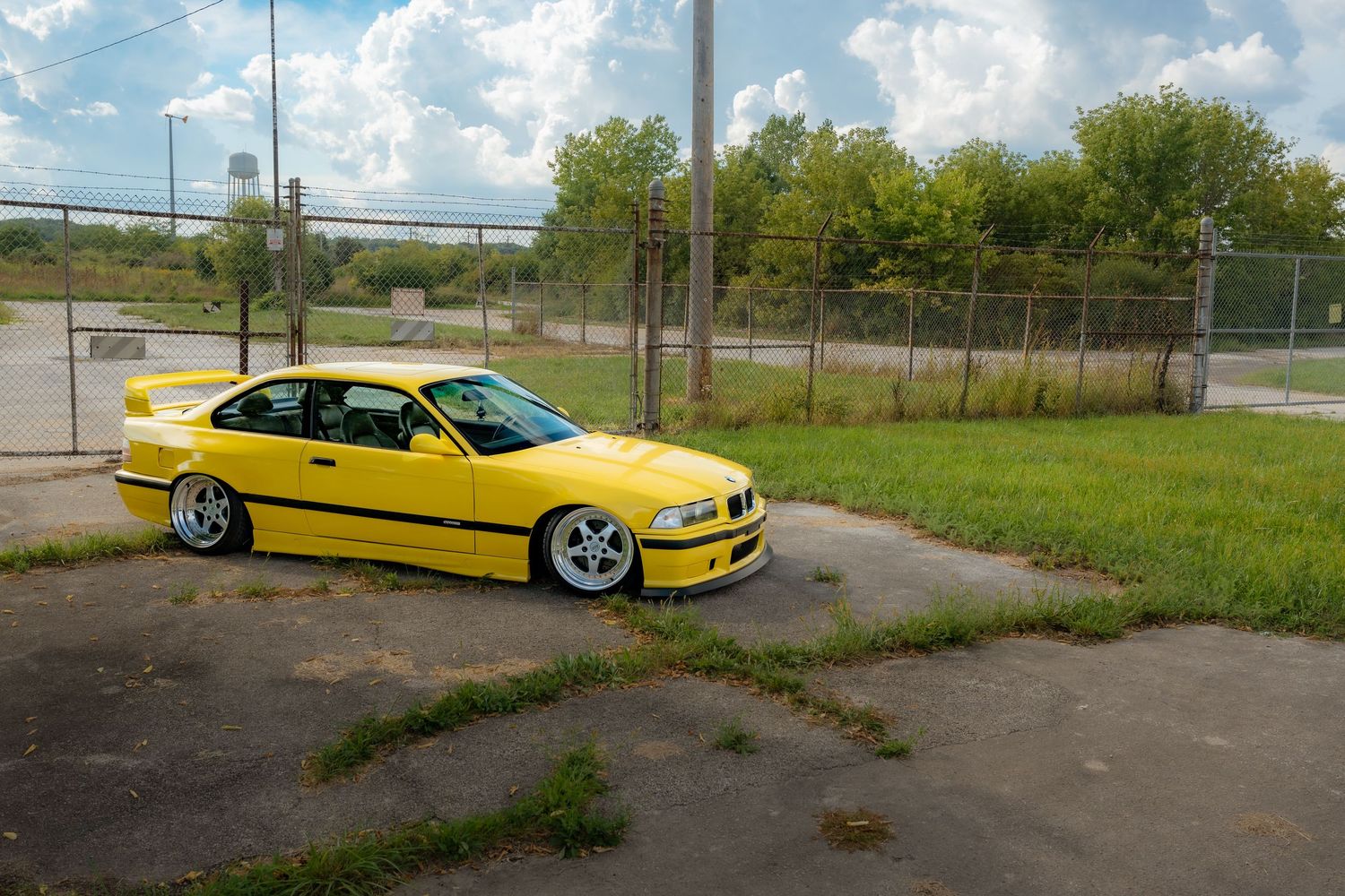 A bright yellow BMW M3 E36 sports car parked in an empty lot showcases its sleek design and aftermarket wheels.