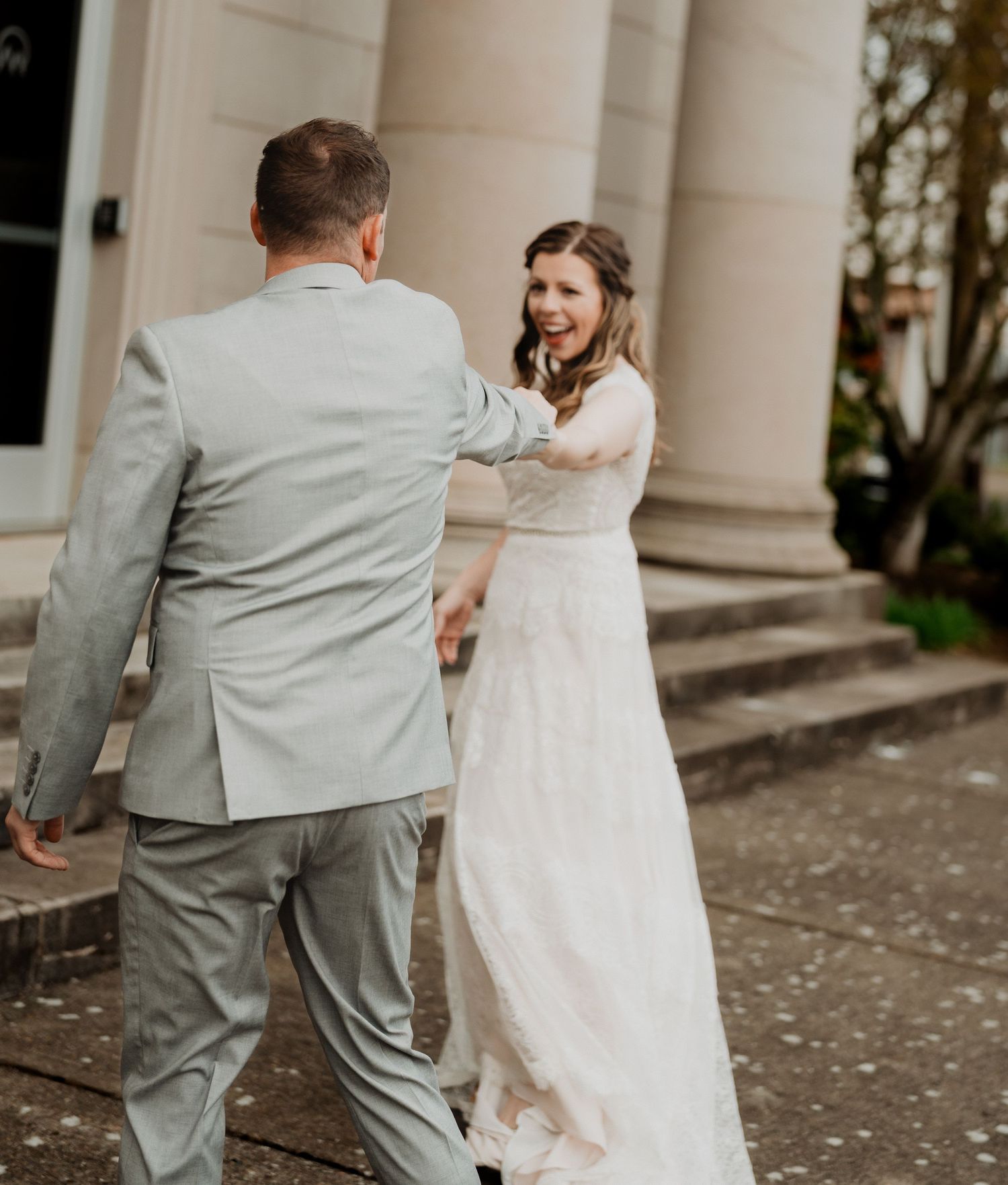 A wedding couple shares a joyful moment on stone steps with the bride in a flowing white dress and groom in light gray suit.